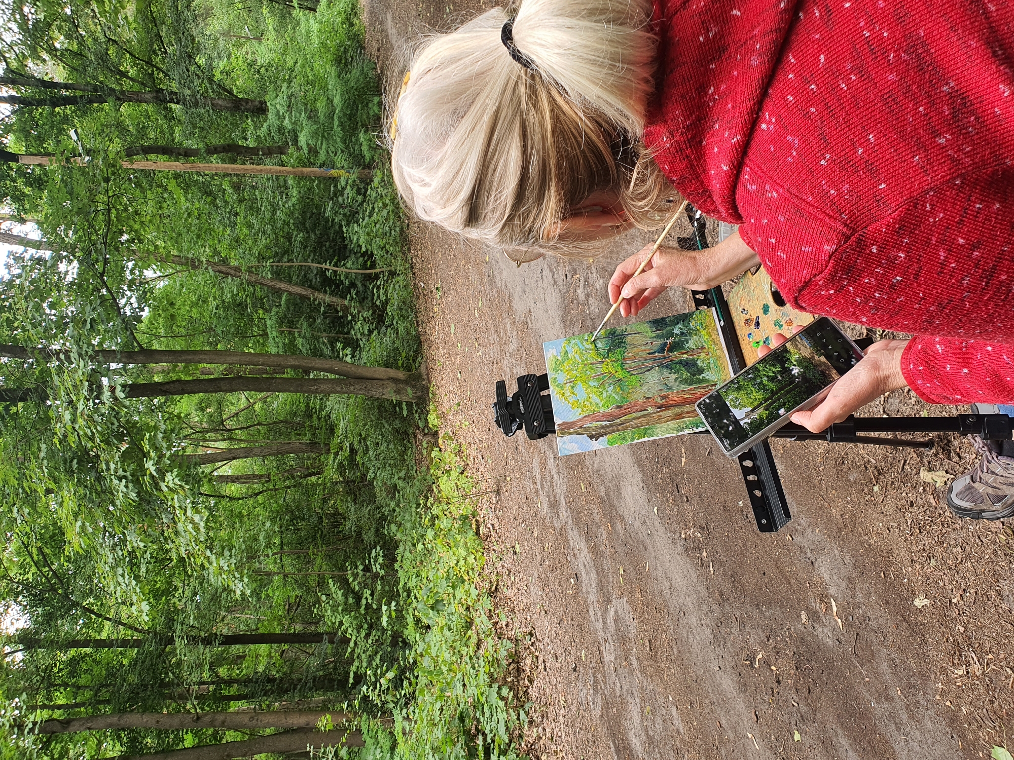 A blonde woman is painting a landscape in a wooded area of Falkensee, Germany. She is wearing a red hooded jacket and is seated, facing away from the viewer, towards a small easel. On the easel is a canvas depicting a forest scene, with trees and foliage. The woman is holding a paintbrush in her right hand and a smartphone in her left hand, which is displaying an image of the same landscape. The smartphone is mounted on a small stand. The ground is covered in dirt and fallen leaves. The trees are tall and green, with dense foliage. The overall impression is one of an artist capturing the beauty of nature.