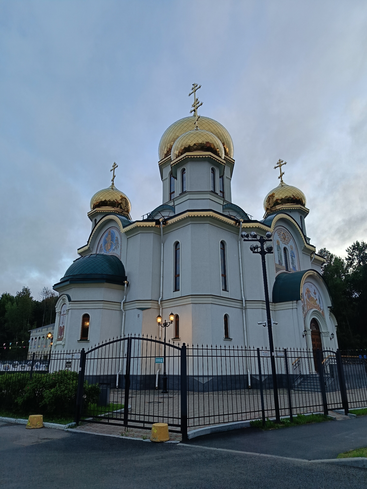 This is a photograph of a white orthodox church with multiple golden domes, each topped with a cross, against a cloudy sky. The church features arched windows and decorative moldings. Mosaic murals are visible on the exterior. A black metal fence with pointed finials encloses the property, and a sign reading "ВХОД" (Entrance) is posted on the gate. The ground in front of the fence is paved, and yellow concrete bollards are visible. To the left of the church, there is landscaping with green bushes and trees in the background. The lighting suggests it is either early morning or late evening.