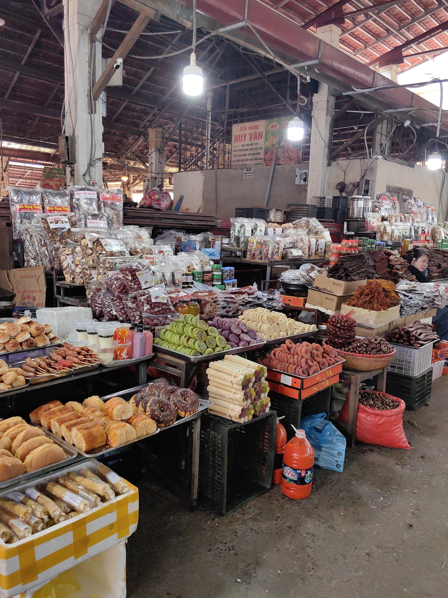 This image captures an indoor market stall in Sa Pa, Vietnam, as indicated by a prominent sign for "CUA HANG HUY VAN" advertising "ĐẶC SẢN SAPA" (Sa Pa specialty). The environment features a high, exposed ceiling with a tiled roof, visible wooden and metal structural beams, and concrete pillars. Multiple bright LED light bulbs hang from the ceiling, illuminating the densely packed scene. The floor is a dark, uneven concrete surface. A white surveillance camera is mounted high on a concrete pillar in the upper left background.
A wide array of food products is displayed on metal tables, shelves, and plastic crates. In the foreground, from left to right, are trays of light-colored rectangular pastries, small golden buns, and chocolate-frosted donuts. Below these, a white polystyrene container with yellow tape holds numerous individually wrapped cylindrical snacks.
The mid-ground is dominated by extensive displays of both packaged and bulk food items. Clear plastic bags containing dried goods such as mushrooms and various fruits, some with Vietnamese labels like "TĂNG TỐC VUA MẸ," are stacked high. Adjacent displays include trays of grilled meat skewers, plastic cups with multi-colored beverages, pink and purple bottled liquids, green circular pastries adorned with kiwi slices, purple and light-colored dried fruit slices, and orange ring-shaped dried persimmons contained in plastic crates. Bulk quantities of reddish-brown dried meat, shredded orange-brown meat floss, and dark round nuts (consistent with chestnuts) are presented in cardboard boxes, woven baskets, and a red fabric bag. Small jars of condiments are interspersed.
A single individual, a female, is partially visible from the head and shoulders in the mid-right, positioned behind a display of dried meats and looking towards the left.
On the floor in the lower right, a large orange plastic bottle, labeled "LaVie," and a blue plastic bag are present. Black and grey plastic crates are utilized throughout the scene for display and storage.