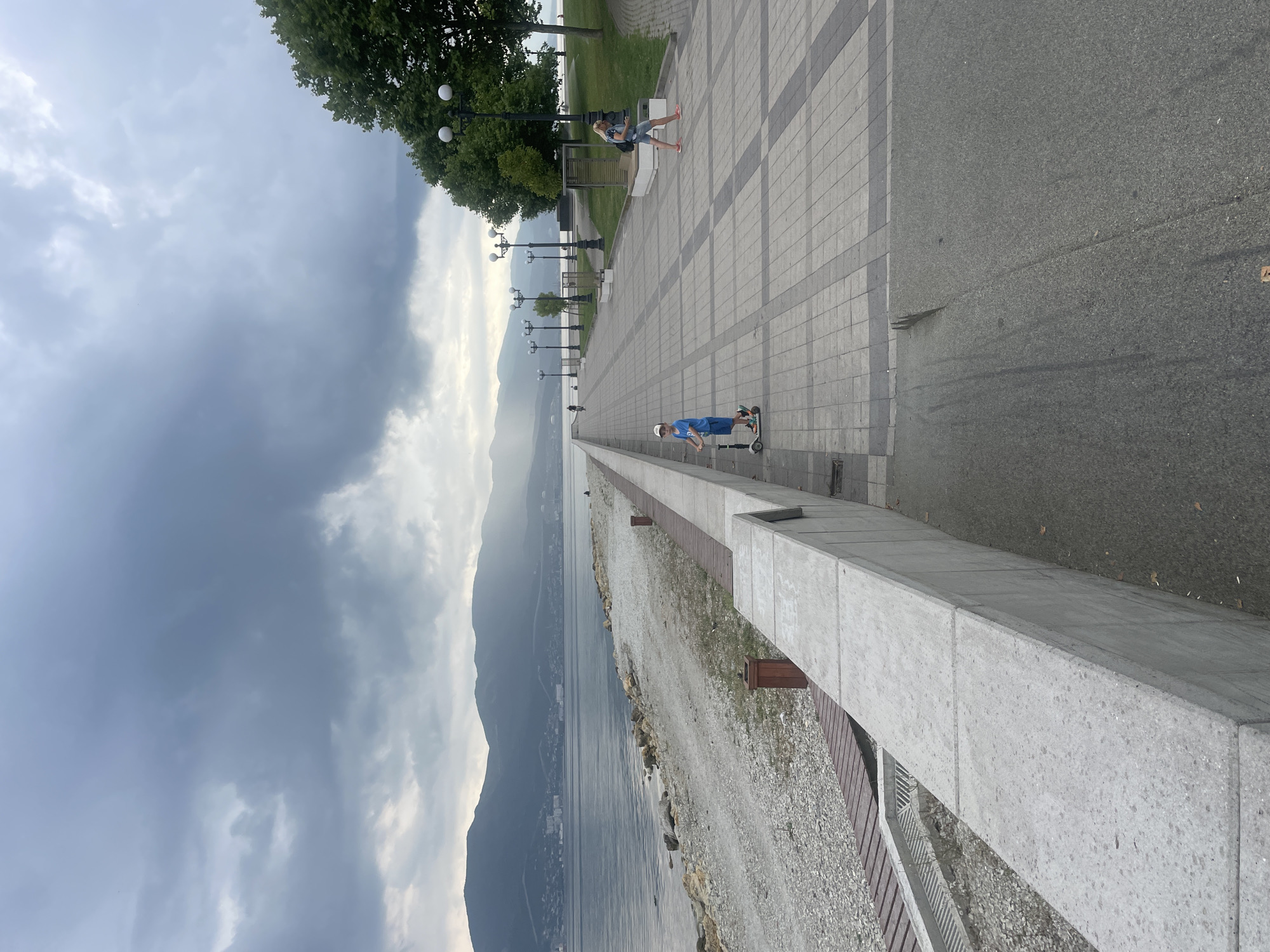 The image depicts a cloudy day at a seaside promenade in Novorossiysk, Russia. A young boy is riding a scooter on the paved walkway. Further down the promenade, a woman with a backpack is walking. The promenade overlooks a pebble beach and the calm sea, with a mountainous landscape in the background. Lampposts line the promenade, and trees are visible on the right side of the image. The sky is overcast with heavy clouds.
