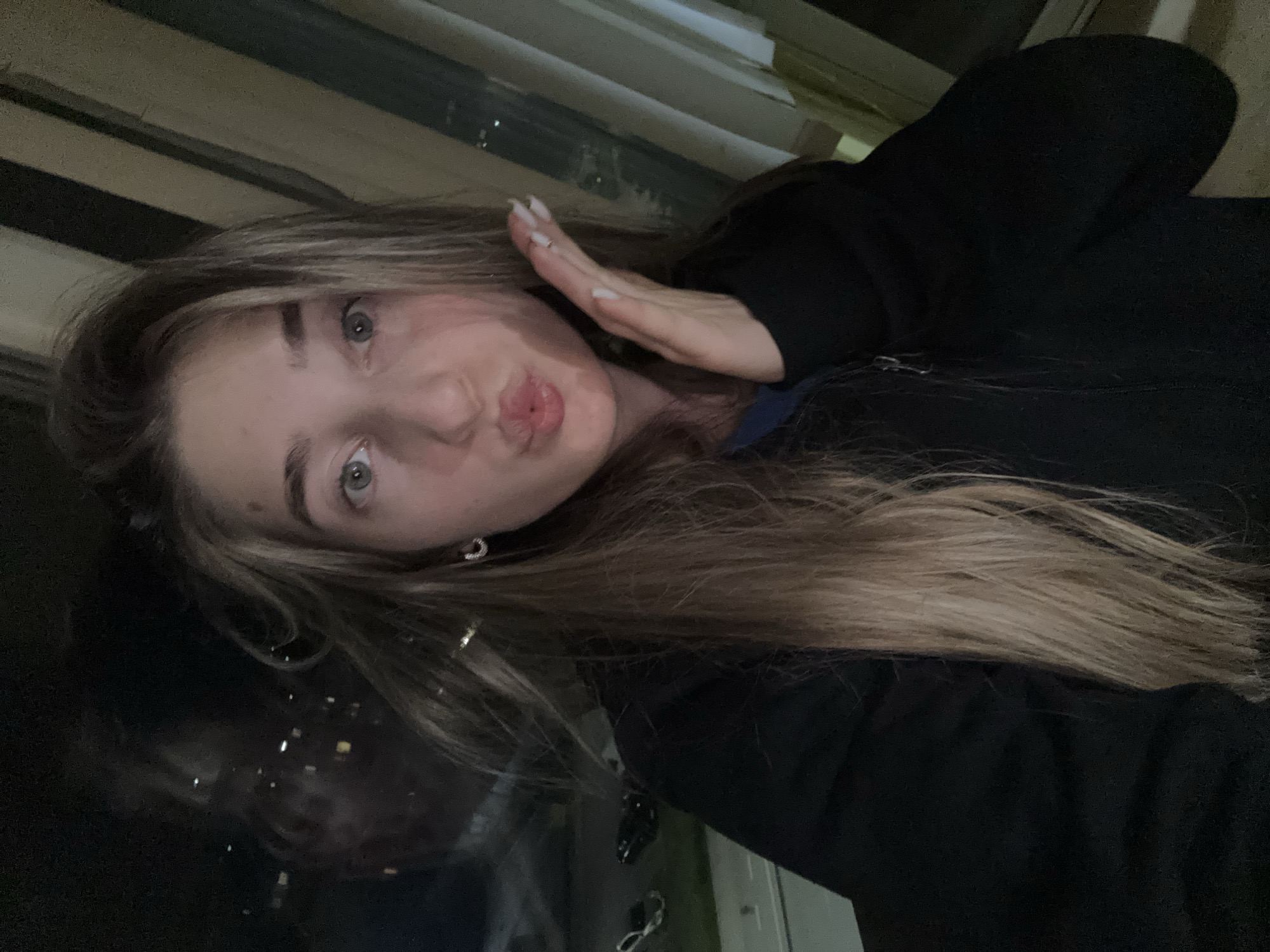 A young woman poses for a selfie with pursed lips and her hand near her cheek. She has long, light brown hair, fair skin, and light eyes. She is wearing a dark-colored sweatshirt. A window is visible behind her, reflecting city lights at night. The location is identified as Unknown, Unknown.