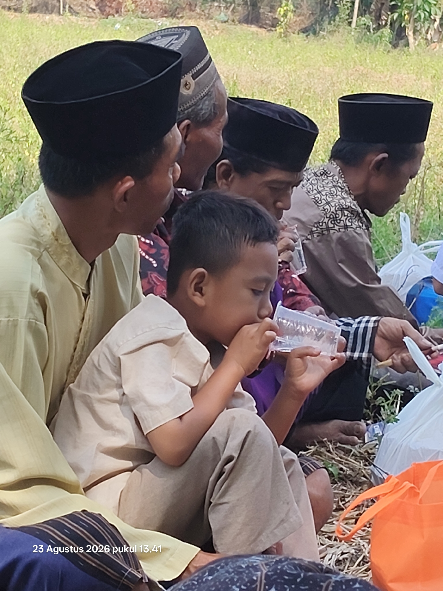 In a sunlit outdoor setting, a group of individuals are gathered, seated on the ground amidst dry grass and vegetation. The scene, located in Padang Mentoyo, Indonesia, suggests a casual communal gathering.
In the foreground, a young boy, wearing a light-colored shirt and pants, is actively drinking from a clear plastic cup held in his hands. His focus is directed towards the cup. To his left, an adult male, dressed in a light yellow garment with embroidered details around the neckline, is partially visible, looking downwards.
Behind them, several other individuals are present. One man wears a dark peci (a type of cap) and is seen in profile, also appearing to drink from a similar plastic cup. Another man, also wearing a peci, has a patterned shirt and is looking down. Further back, another person with a peci is visible.
The environment consists of dry, straw-like ground cover with some green vegetation interspersed. Bags, including a white plastic bag and a bright orange reusable bag, are scattered around, indicating provisions or belongings. The overall atmosphere appears relaxed and informal, with people engaged in quiet activity. The timestamp in the bottom left corner indicates the image was taken on August 23, 2026, at 13:41.