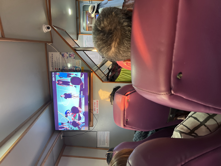 This image, taken inside what appears to be a passenger cabin on a bus or ferry in Hà Tiên, Vietnam, shows several people seated, primarily from behind. The seating consists of individual purple-colored, upholstered chairs, arranged in rows.

In the foreground, the top of a person's head, with graying hair, is visible. Beyond them, the back of another person's head is seen, also in a purple seat. Further back, another person's head is visible, wearing what looks like a white brimmed hat.

The main activity is watching television. A large flat-screen TV is mounted on the wall to the left (when viewing the image upright). The screen displays what appears to be a sports event or a performance involving people on a court, possibly playing basketball or dancing, with several figures in athletic wear. The text "SO PRESS" is visible on the TV screen, along with some smaller, unreadable Vietnamese characters, likely subtitles or channel information.

Notable details include a white security camera (CCTV) mounted on the ceiling in the upper left. Various informational signs and posters are affixed to the walls. One prominent sign above the TV has Vietnamese text that reads "BẢNG HIỆU QUY ĐỊNH ĐỐI VỚI HÀNH KHÁCH" (Regulations for Passengers). Another sign near the bottom right of the TV clearly displays "ĐƯỜNG DÂY NÓNG" (Hotline) and a phone number "0915.666.660". The lighting suggests an interior space, likely artificial, possibly combined with natural light from unseen windows. The overall setting is functional, typical of public transport.