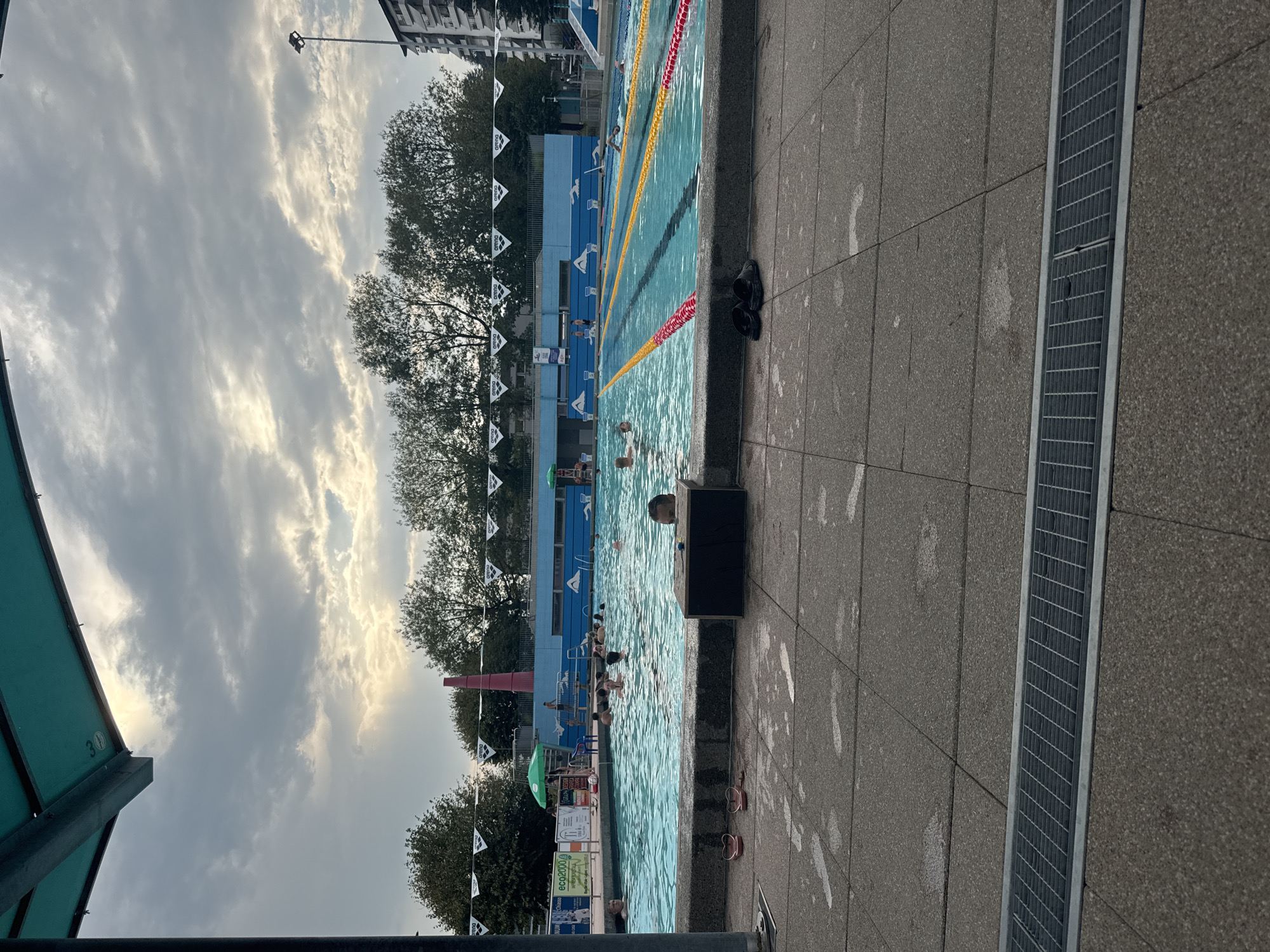 The image depicts an outdoor swimming pool facility on an overcast day, likely located in Chiasso, Switzerland. In the foreground, a grey tiled patio area with visible water marks and a metal drainage grate runs along the bottom edge. Two pairs of black flip-flops are placed on the patio. Beyond the patio is the swimming pool, divided into lanes by yellow and red buoys. Several people are in the water, some swimming and others standing or sitting. In the background, trees line the edge of the pool area, with a blue building and apartment blocks visible beyond the trees. A tall lamppost stands to the right of the apartment blocks. Triangular "arena" flags are strung along the top edge of the pool area. To the left, a green umbrella shades a lifeguard chair. A red chimney-like structure is visible behind the trees on the left side of the image.