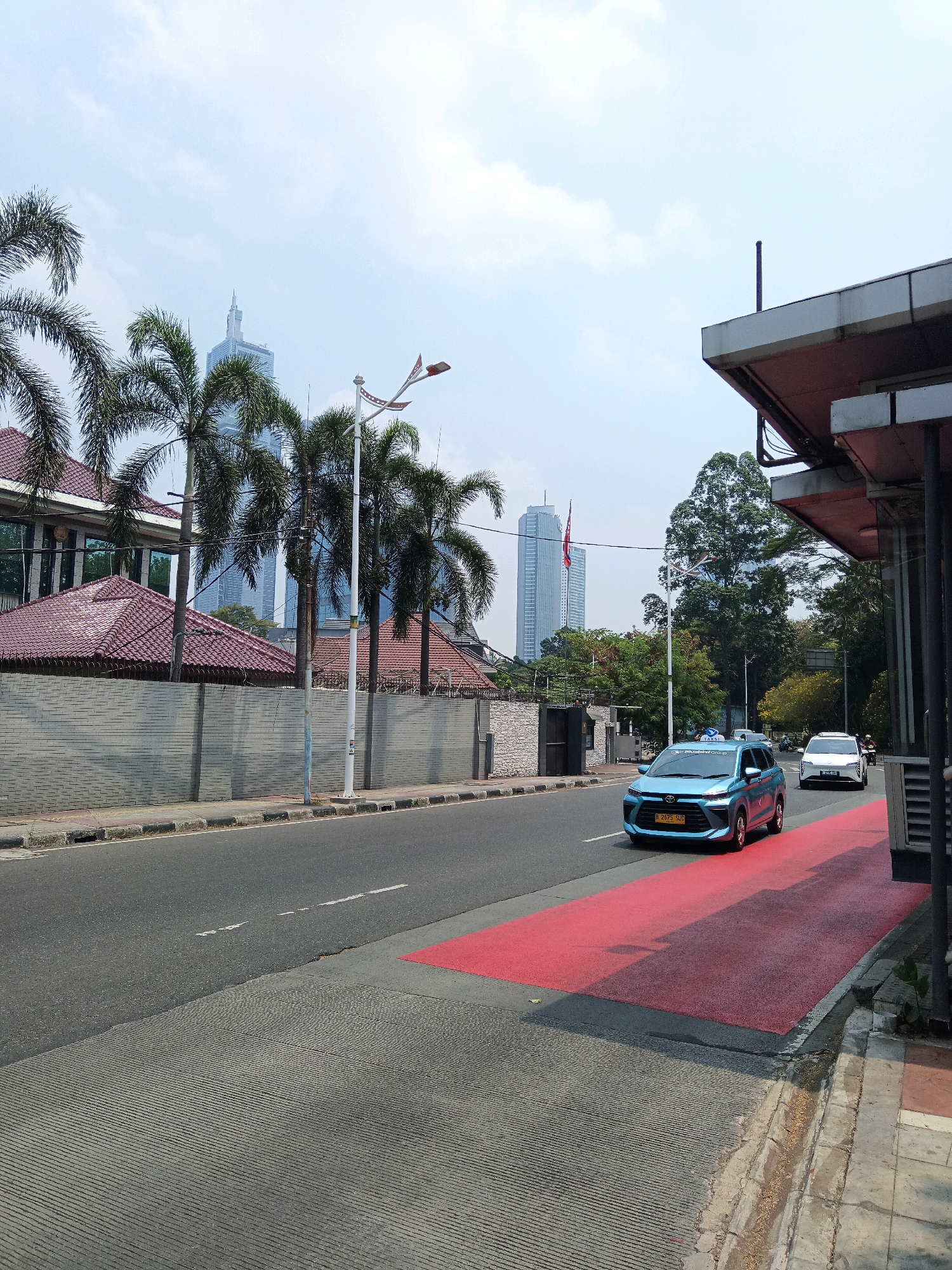 The image depicts a street scene in South Jakarta, Indonesia, likely during daytime hours, given the bright sky and clear visibility. In the foreground, a light blue taxi with license plate "B 2075 SUP" is driving on the road. To the right of the taxi is a red-painted area on the road, possibly indicating a pedestrian crossing or a special lane. A white SUV is visible further down the road, also in motion.
The background features several tall buildings, one of which is a prominent skyscraper with a distinct spire. Palm trees are scattered throughout the scene, and a wall with a patterned design borders the property on the left side of the road. A building with a reddish-brown tiled roof is visible behind the wall, partially obscured by the palm trees. A street lamp with a decorative element, possibly a flag or banner, stands near the palm trees. The environment appears urban, with a clear sky overhead and some clouds. The presence of the taxi and the modern buildings suggests a well-developed urban area. The infrastructure, including the road, sidewalk, and drainage system, is visible. The overall impression is that of a typical city street with vehicular traffic and architectural elements characteristic of South Jakarta.