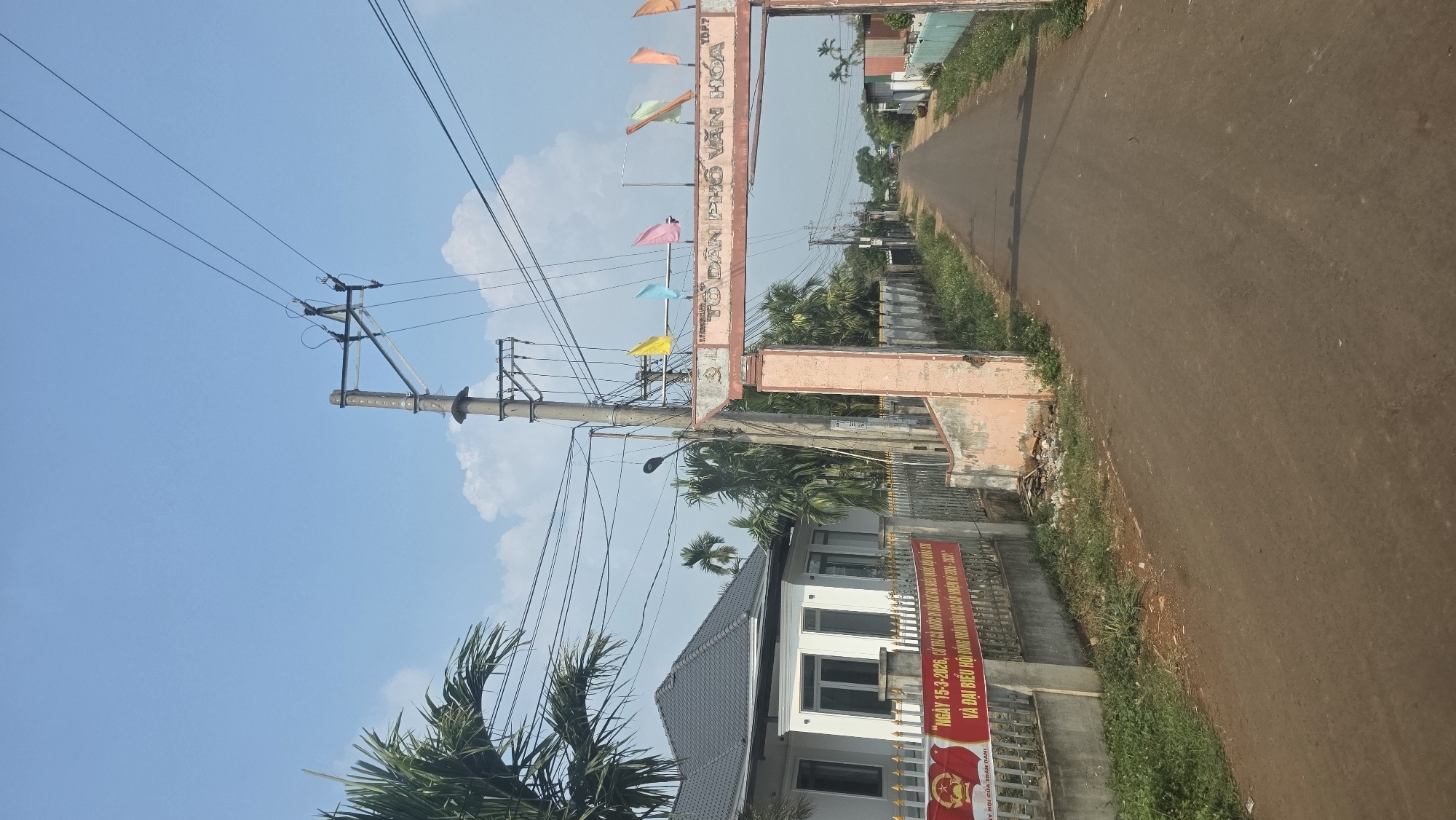 The image depicts a street scene in Xuân Long, Vietnam. A road stretches into the distance, bordered by vegetation and structures. An ornate archway with colorful flags spans the road, bearing text in Vietnamese. To the left, a utility pole with power lines stands prominently. A two-story building with a gray tiled roof and white walls is visible. A banner with red and gold colors, also with Vietnamese text, is attached to the building's fence. The sky is blue with scattered clouds, and palm trees are visible in the foreground.