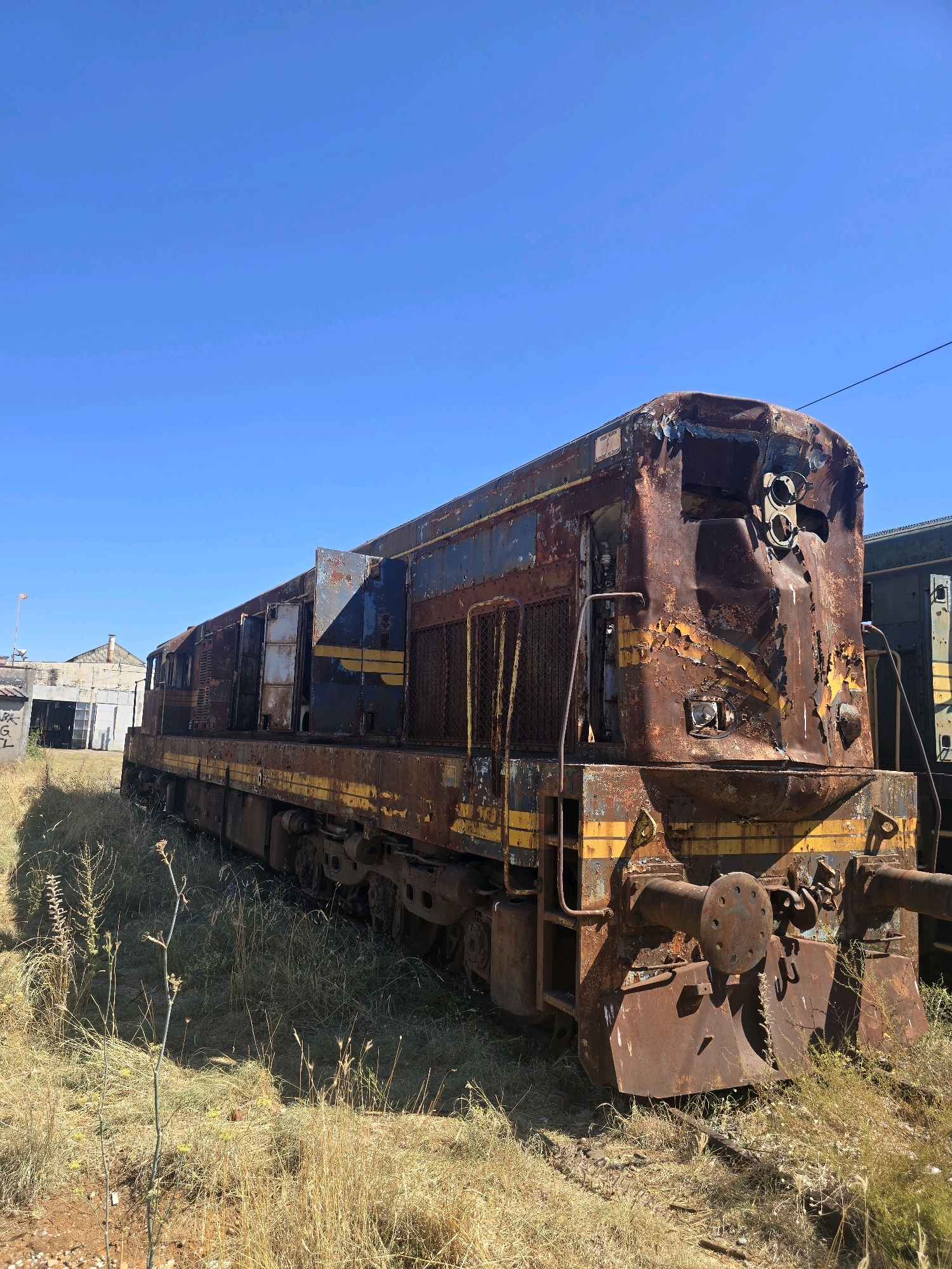 The image shows a heavily rusted, abandoned train engine parked on a dry, grassy field under a clear blue sky. The engine is predominantly brown with streaks of faded blue and yellow paint. It appears to be a diesel locomotive, with visible features such as a large coupling mechanism at the front, wheels, and windows. In the background, a light-colored building with a corrugated roof is visible, along with some trees and a power line. The area appears to be an industrial or train yard setting. The overall condition of the train suggests it has been decommissioned and left to decay for a significant period. There are no people or animals present in the image. The scene conveys a sense of desolation and the passage of time.
