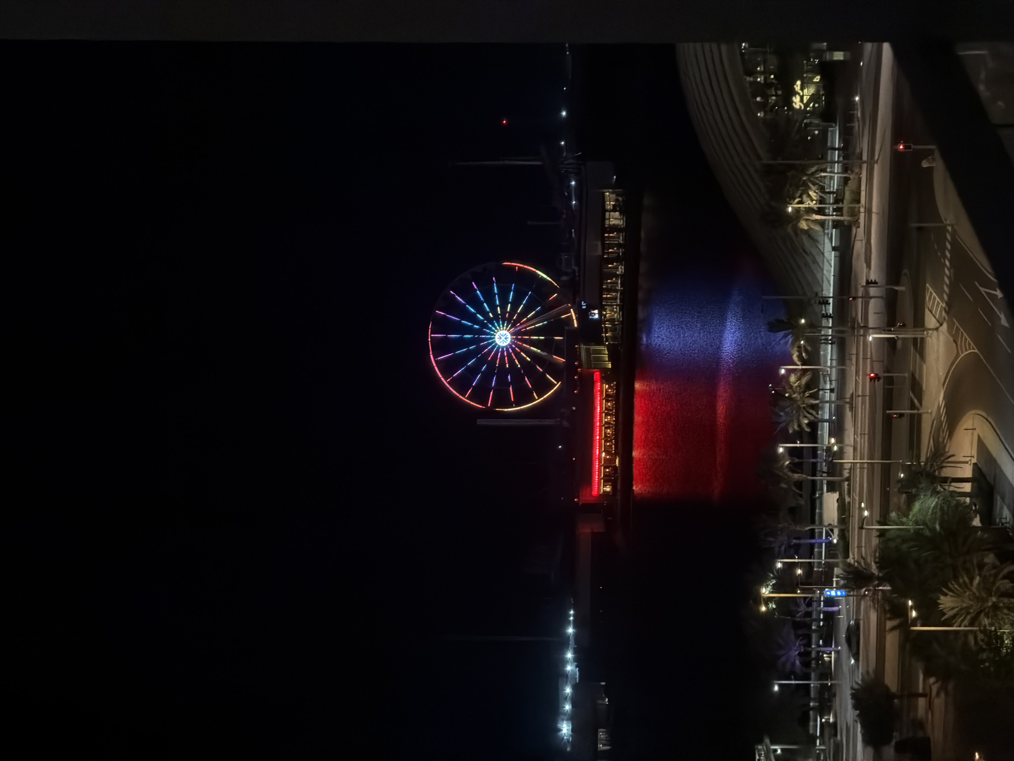 The image depicts a nighttime scene in a city. A large Ferris wheel illuminated with colorful lights is the central focus, situated across a body of water. The Ferris wheel is adorned with red, yellow, blue, and purple lights, casting reflections on the water. Adjacent to the Ferris wheel, a building with red and white lights is visible, possibly a restaurant or entertainment venue. The water itself reflects the colorful lights from the Ferris wheel and the building.
In the foreground, a street scene is depicted with palm trees lining the road. Streetlights illuminate the area, and traffic signals are visible. A car is parked on the side of the road, and a white van is also present. Beyond the street, a large, curved architectural structure with lights can be seen, suggesting a modern building or complex. The overall environment suggests an urban area with recreational and commercial spaces, likely a waterfront district. The image is framed by a dark structure on the right side, possibly a balcony or window.