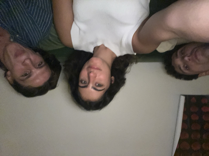 Three young adults lie close together, their faces near the camera, creating a selfie-style image. On the left, a person with short dark hair wears a striped shirt. The individual in the middle has long, dark, wavy hair and a white top. To the right, another person with short, curly hair is wearing a beige shirt. They appear to be lying on a green surface or bed. Part of a patterned item, possibly a book or painting, is visible in the lower right corner.