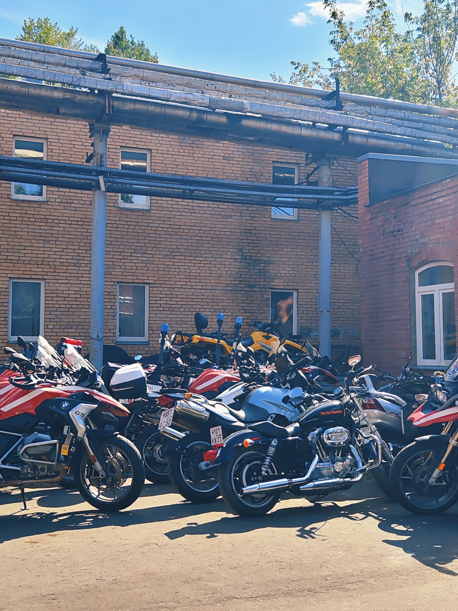 A sunny outdoor scene depicts a row of parked motorcycles in front of a brick building. The motorcycles are varied in color and style, with some featuring red and white detailing and others in darker colors. They are positioned on an asphalt surface, and the building behind them has several windows. Overhead, large insulated pipes are supported by a metal structure. The sky is blue with some white clouds. The overall impression is that of a parking area for motorcycles in an urban industrial or commercial setting.