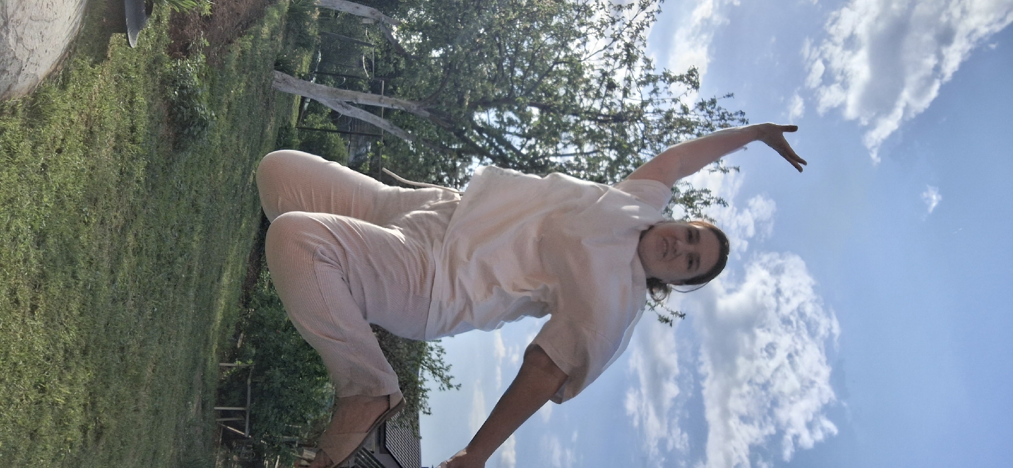 A person is photographed outdoors under a blue, cloudy sky. The person is wearing light-colored clothing (likely pants and a shirt) and sandals. They appear to be kneeling or crouching on a surface, with one arm raised. The background includes green grass, trees, and what appears to be a white wooden structure or fence. The location is in Chaikovsky, Russia.