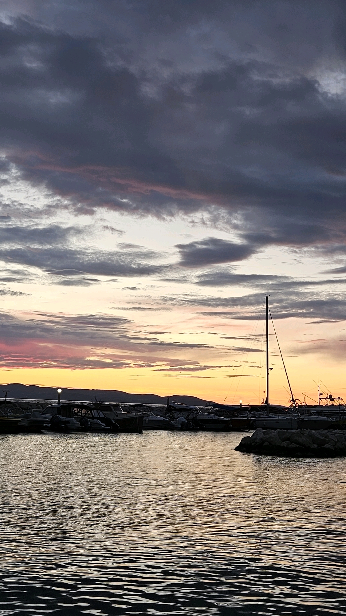 The image depicts a sunset over a harbor in Općina Tučepi, Croatia. The sky is filled with dramatic clouds, ranging in color from dark grays to soft pinks and oranges, illuminated by the setting sun which is visible as a small, bright disc on the horizon. The water in the foreground is rippled, reflecting the colors of the sky. A marina is visible in the middle ground, with several boats docked. One sailboat with a tall mast stands out against the sky. In the background, the silhouette of hills or mountains can be seen. The overall scene conveys a sense of calm and natural beauty.