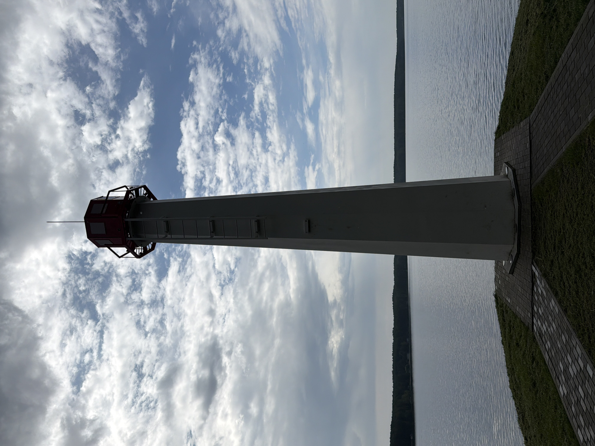 A tall, white lighthouse stands on a grassy knoll overlooking a calm body of water. The lighthouse has a red top with glass windows and a metal railing. A ladder is attached to the side of the lighthouse. The water stretches out to a tree-lined horizon under a cloudy sky with patches of blue. Paved pathways lead away from the base of the lighthouse across the grass. This lighthouse is located in Zarechny, Russia.