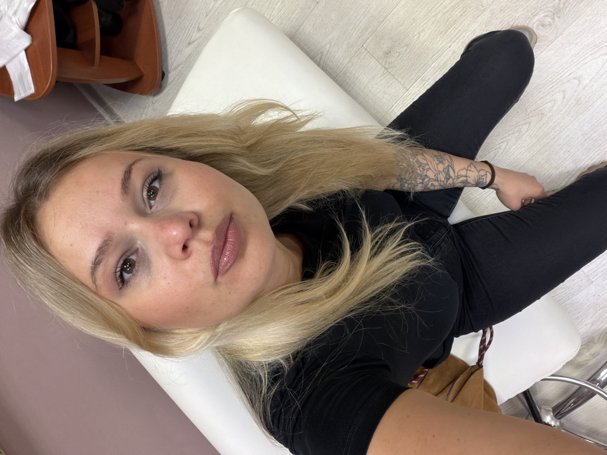 A blonde woman with fair skin, clad in a black t-shirt and black jeans, is positioned on a white padded chair. A brown suede handbag rests on the chair beside her. The woman has a tattoo on her left forearm. The location appears to be indoors, possibly a salon or a studio setting. The flooring is light-colored wood-like or laminate, and other furniture, including a shoe rack, is partially visible in the background. There is no indication that this image was taken in the city of Sarapul, Russia.