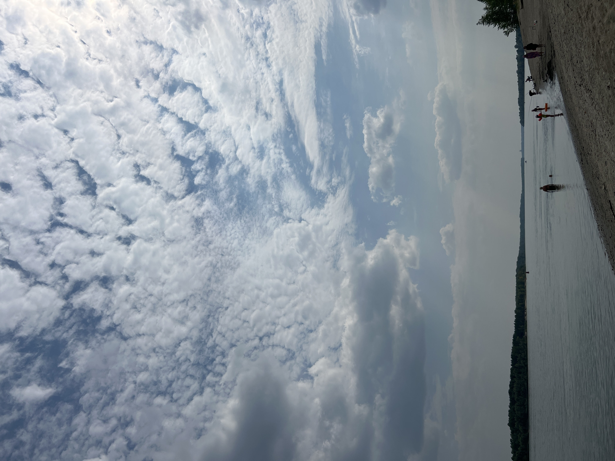 The image depicts a beach scene along a wide river, possibly the River in Mochishche, Russia. The sky is filled with a variety of clouds, ranging from dark, heavy formations on the left to lighter, scattered clouds with patches of blue sky on the right. The sun is visible through the clouds, casting a bright light.
On the sandy beach, several individuals are present. Some are in the shallow water, while others are on the shore. One person is standing in the water, and another appears to be swimming. Further down the beach, a group of people are gathered, with some appearing to be walking or standing. One individual is holding a bright orange flotation device. The sand on the beach is a light brown color, and there are some footprints visible.
Across the river, a dense line of trees forms the shoreline of the opposite bank, stretching into the distance. A red buoy is visible in the water near the opposite shore. The water itself appears calm, reflecting the sky. The overall impression is a serene, outdoor scene with people enjoying the river and beach on a cloudy but bright day.