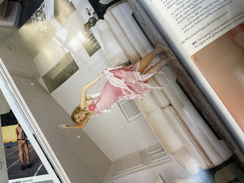 The image displays an open magazine spread, photographed at an oblique angle. The primary subject is a full-page color photograph on the right, depicting a fair-skinned woman with long, blonde hair, smiling with closed eyes, captured in a dynamic pose as if dancing or twirling. She wears a sleeveless, reddish-pink dress with a white checkered pattern, ruffled detailing, an asymmetrical hemline, and fringes. A pink floral embellishment is visible on her left shoulder strap. Her left arm is raised above her head with an open hand, and her right arm is bent, seemingly adjusting her dress. She is barefoot.
The scene is an interior space, likely a bright, modern living area with white or off-white walls and a high ceiling. A long, light-colored (beige or off-white) three-seater sofa with distinct seam lines and wide armrests dominates the lower part of the photograph. In the upper right background, multiple rectangular window-like openings or architectural cutouts feature decorative white ironwork grilles. On the left wall, near the floor, a set of white panel doors or cabinets is visible. A dark, ornate side table is positioned behind the right end of the sofa, holding a vase with a colorful floral arrangement and other decorative items. Recessed lighting is visible in the ceiling. The floor appears to be a light, possibly grey or stone surface, showing some reflections.
On the upper left page of the magazine, a fragment of text "football case study" is visible alongside a photograph of a man in a light brown suit lying on a paved surface. Text from an interview, including labels "YD:" and "BH:", is visible on the lower right page of the magazine.
The depicted indoor scene offers no visual identifiers or landmarks to specify a location within Paris, France, or confirm the city itself.