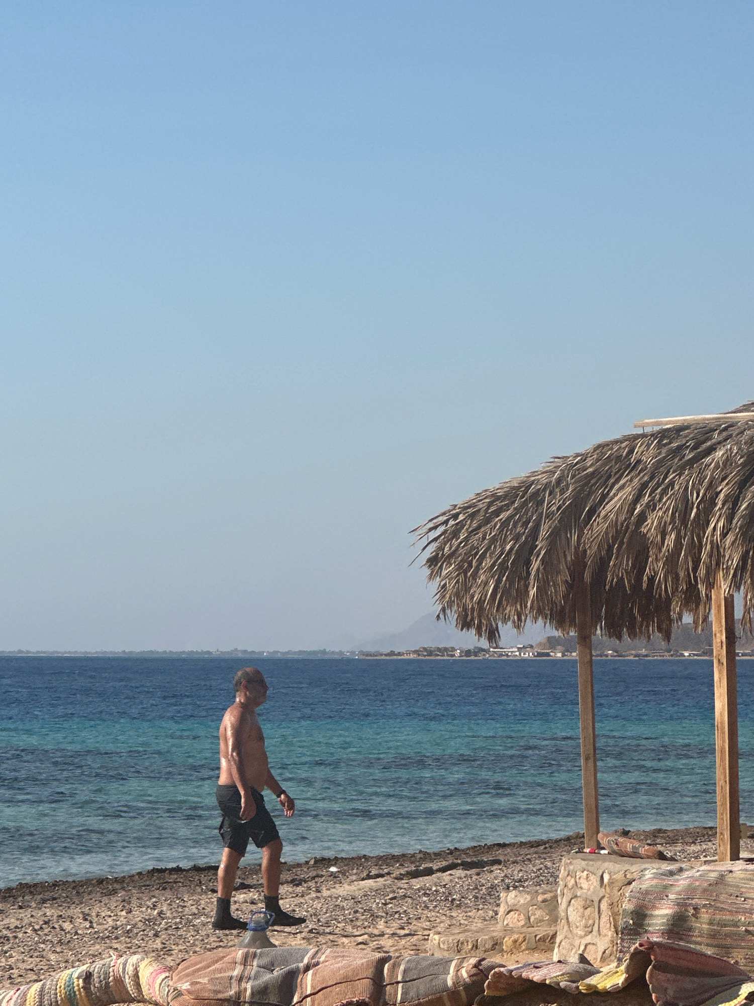 The image depicts a beach scene in Nuweiba, Egypt. A man, appearing to be in his late 50s or early 60s, walks along the shore. He is wearing dark shorts and black socks. His skin is glistening, suggesting he may have recently been in the water or is sweating. The man is positioned on a pebble beach adjacent to the turquoise waters of the sea, which stretch out to a distant coastline marked by hills. A thatched umbrella, supported by wooden poles, provides shade on the right side of the frame. In the foreground, colorful textiles are spread out on the ground, suggesting a resting area. The sky is clear and blue.