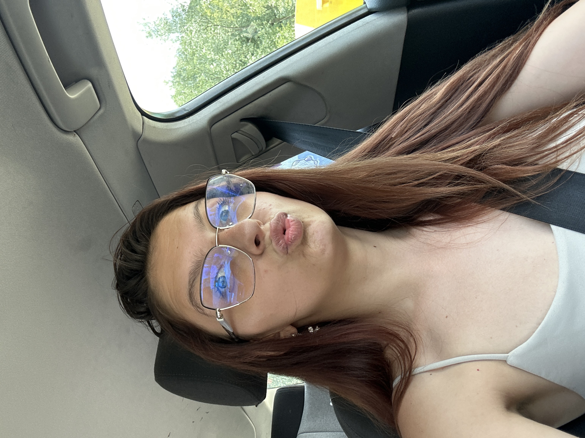 A young woman with long, reddish-brown hair, wearing glasses and a light-colored tank top, is sitting in the driver's seat of a car. She is looking forward and has her lips puckered. The interior of the car is visible, including a seatbelt, headrest, and part of the dashboard. Outside the car window, trees and a bright yellow object are visible. The scene appears to be in a natural setting, possibly on the outskirts of Tryavna, Bulgaria, indicated by the lush greenery.
