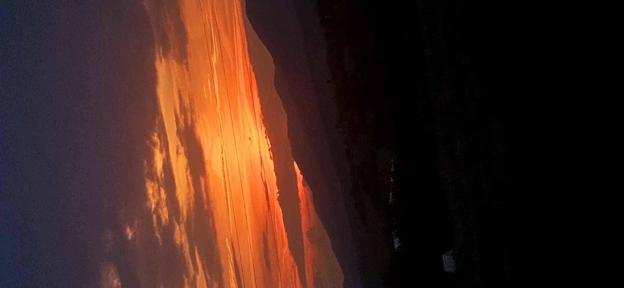 The image displays a mountainous landscape at sunset or sunrise, located in Momin Prohod, Bulgaria. The sky transitions from a deep indigo at the top to vibrant orange and red hues towards the horizon, where the sun's light is most intense. Numerous clouds are present, illuminated from below in shades of orange and red, forming horizontal streaks and clusters. Faint, thin lines, consistent with overhead wires, traverse the mid-sky horizontally. Below the horizon, successive layers of mountains or hills are visible as dark silhouettes against the bright sky, creating a sense of depth. A small, intense red glow is discernible in a valley between the closer mountain ranges. The immediate foreground is heavily shadowed and appears as a dark, vegetated area. On the far left of the foreground, a small structure with a light-colored, pitched roof is partially visible. No people or specific actions are depicted.