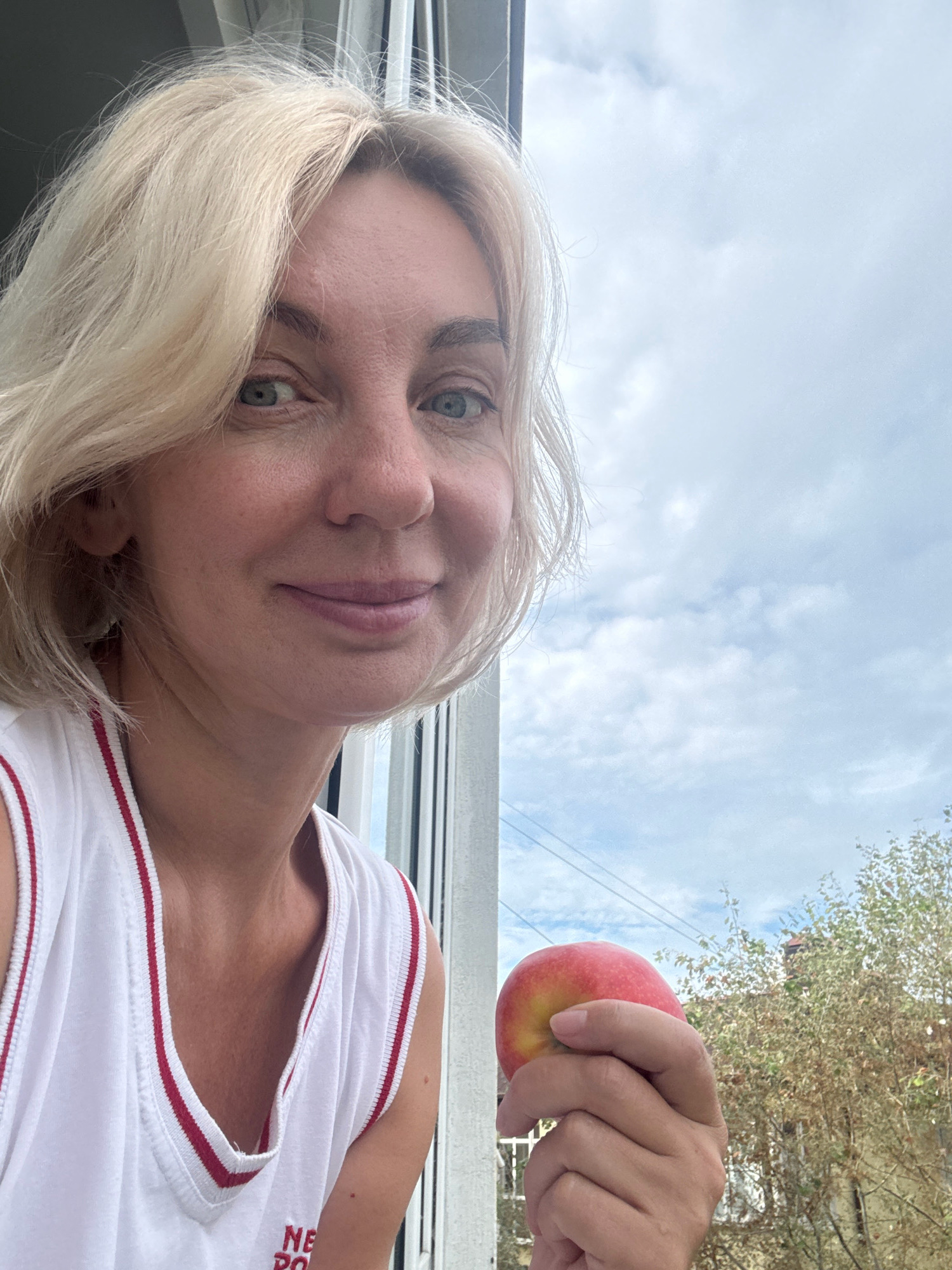 A woman with blonde bobbed hair and green eyes, wearing a white tank top with red trim, is holding a red and yellow apple in her right hand. She is looking towards the camera with a slight smile. The background shows a cloudy sky and some trees. The image appears to be taken from a window or balcony.