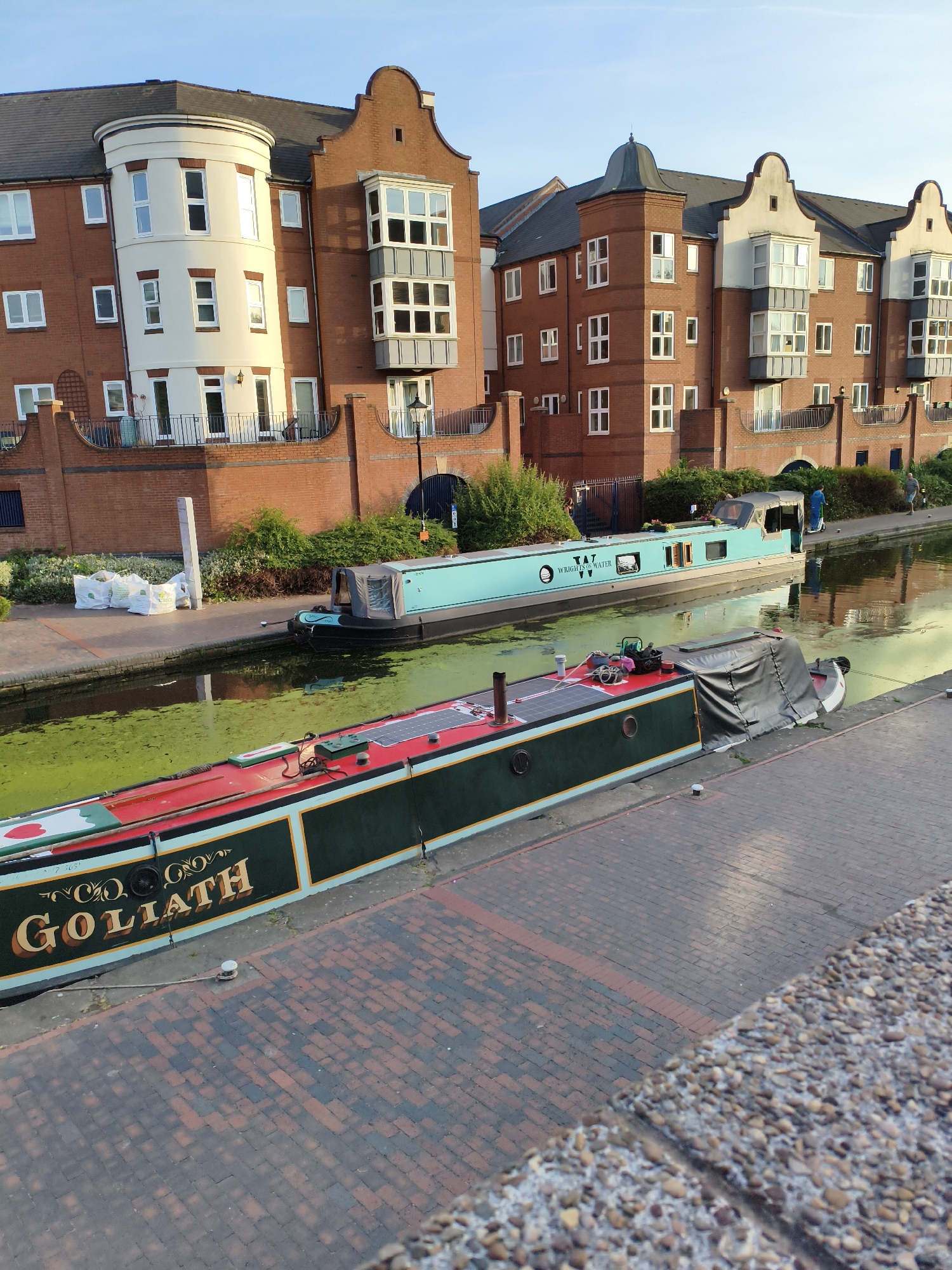 The image captures a scene along a canal in Birmingham, United Kingdom, likely near the city center given the modern apartment buildings. In the foreground, a narrowboat named "GOLIATH" is moored, distinguished by its dark green hull with red and gold trim. Solar panels are visible on its roof, suggesting a degree of self-sufficiency. The water of the canal is covered with a thick layer of green algae. Behind "GOLIATH", another narrowboat, light blue in color, is also moored. This boat has "TAT WRIGHLS & WATER" printed on its side.
The background features a row of contemporary brick apartment buildings with distinctive architectural elements, including rounded turrets and bay windows. These buildings suggest a redeveloped urban waterfront area. Along the canal bank, there is a paved walkway made of red bricks. Greenery and bushes line the canal, interspersed with a few white bags of soil. In the distance, a few people can be seen walking along the towpath, including one individual in a blue uniform and another in casual clothing. The sky is clear and blue, indicating a bright, sunny day, and the setting sun casts a warm light on the buildings. The overall impression is one of a peaceful urban waterway, blending residential development with the historic charm of canal life.