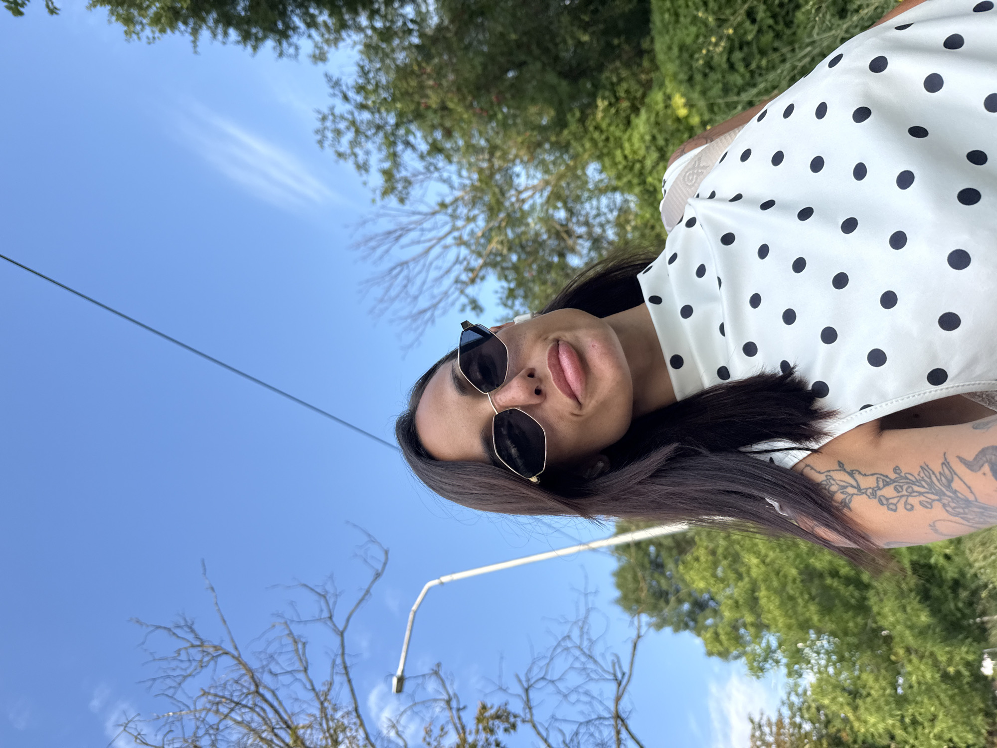 A woman is taking a selfie outdoors. She has dark hair, sunglasses with dark lenses and a gold frame, and is wearing a white top with black polka dots. She has a tattoo on her left arm. She is standing in front of trees and a clear blue sky. A lamppost is visible in the background on the left. The image was taken in Pleven, Bulgaria.