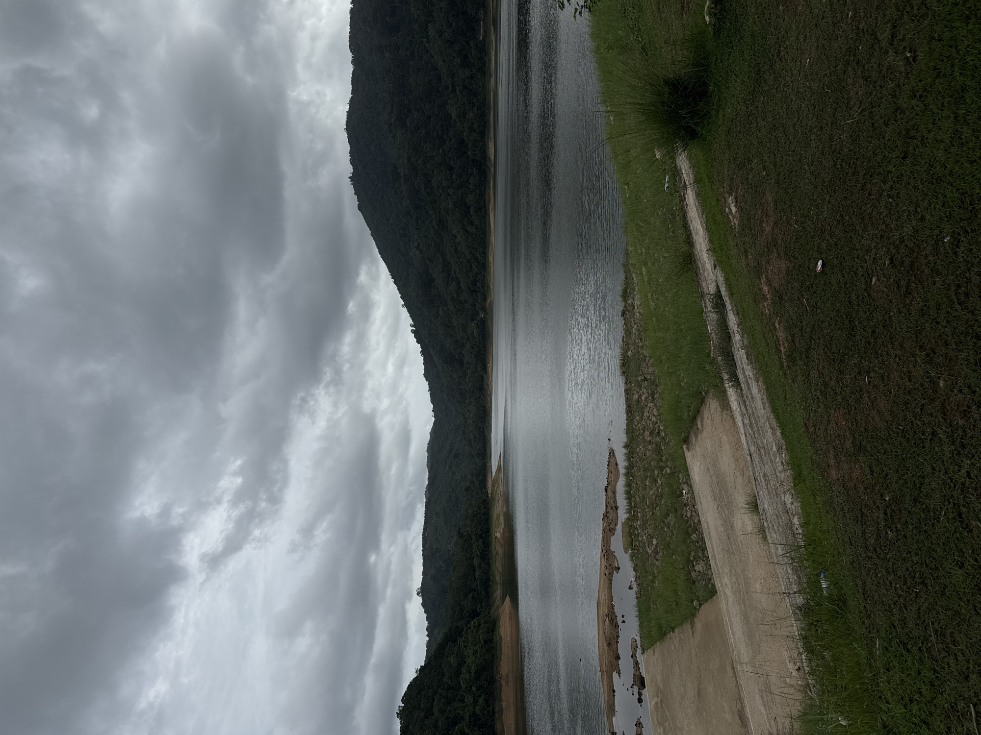 The image depicts a reservoir in Kathu, Thailand under a cloudy sky. A densely forested mountain range borders one side of the water. The opposite side of the reservoir has a grassy bank, with a concrete structure or walkway leading towards the water. The water level appears to be low, revealing a muddy or sandy shoreline. No people are visible in the image.