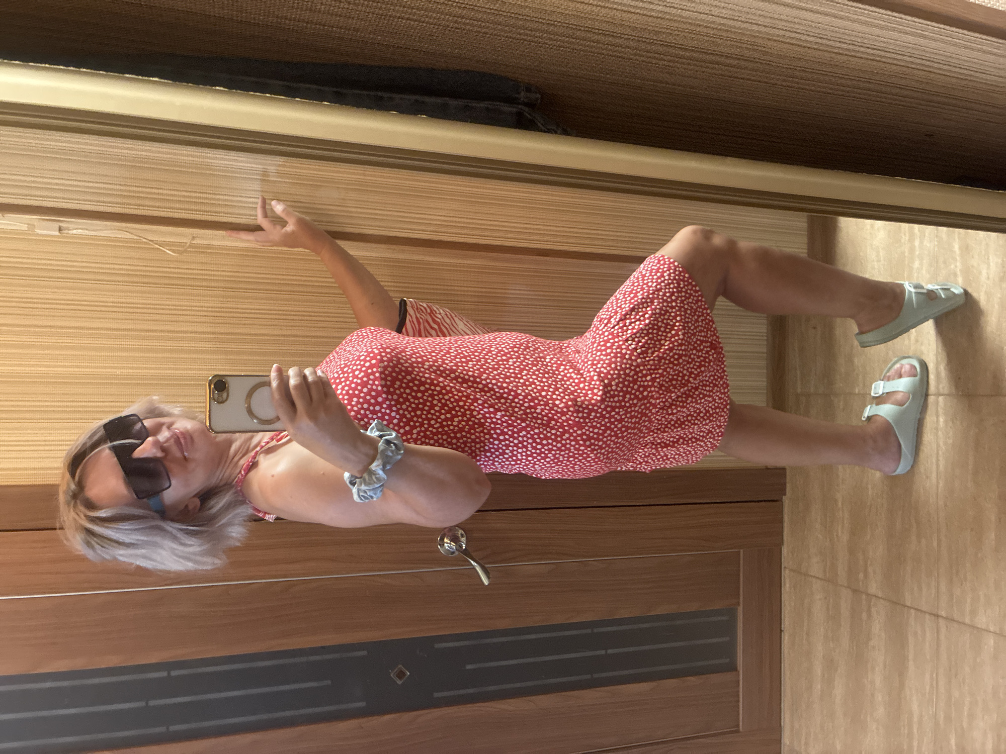 A blonde woman is standing in front of a mirror and taking a selfie. She is wearing a red and white polka dot dress, large dark sunglasses, and light blue slide sandals. She has a scrunchie on her left wrist. The background appears to be an interior, possibly a hallway or closet, with wooden doors and walls covered in vertical blinds. The floor is tiled. The image was taken in Sochi, Russia.