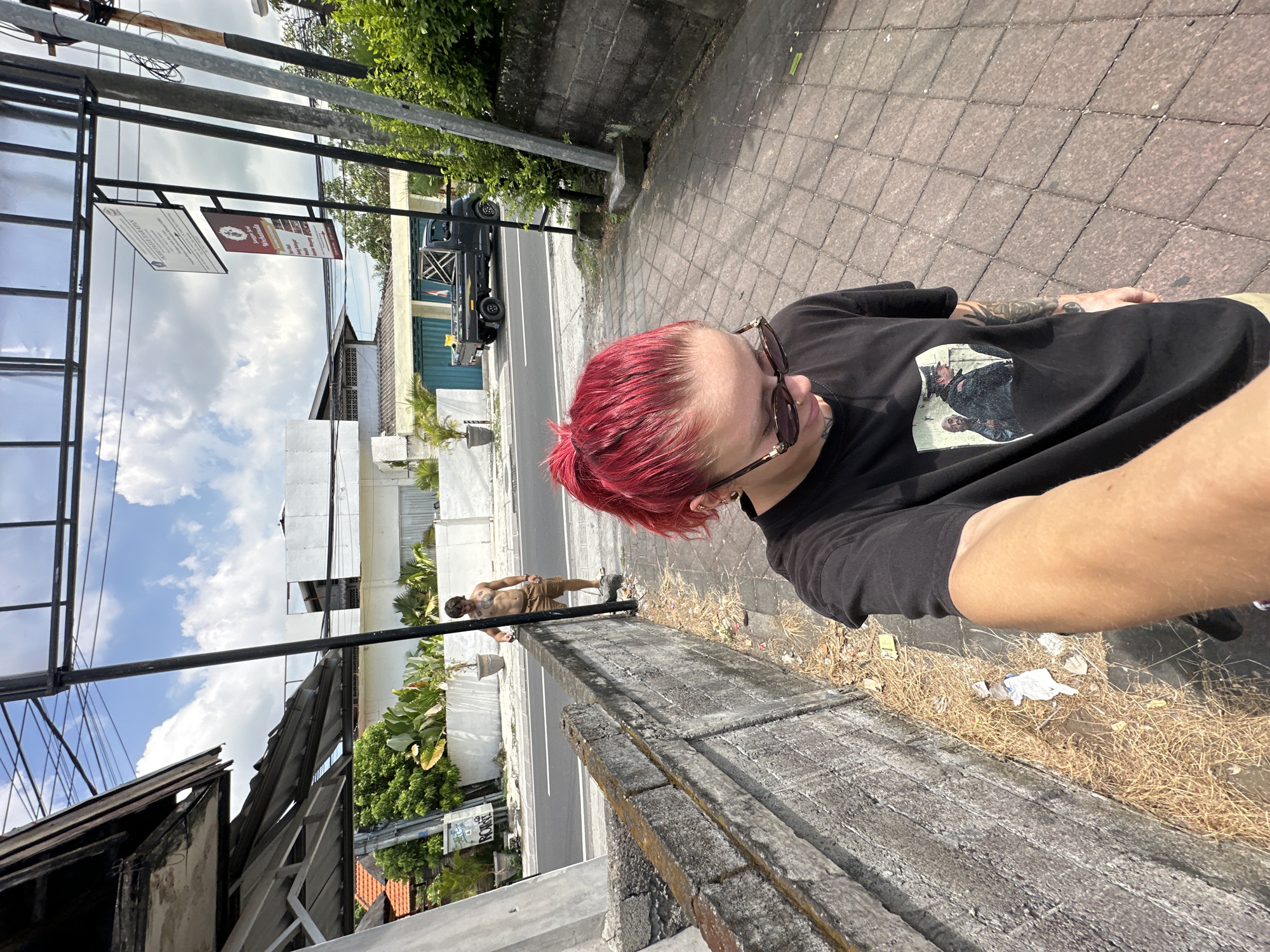 A first-person, medium-distance, angled daytime outdoor shot captures a person with bright pink hair and sunglasses, wearing a black t-shirt with a graphic print. The individual is standing on a paved sidewalk next to a concrete wall. In the background, across the street, stands a shirtless man wearing khaki shorts with his hands on a horizontal metal bar. Further in the background is a road with vegetation on either side, buildings, parked cars, and a cloudy sky. Debris litters the edge of the sidewalk. The location is North Kuta, Indonesia.