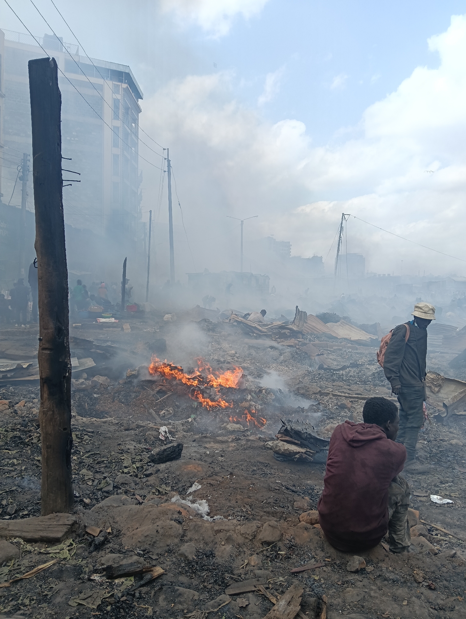 The image depicts the aftermath of a fire, likely in an informal settlement, given the makeshift structures and debris. The scene is heavily obscured by smoke, suggesting the fire has recently occurred or is still smoldering.
In the foreground, two individuals are visible. One, wearing a dark jacket and a light-colored hat, stands with a backpack slung over his shoulder, looking towards the right of the frame. Another person, crouched on the ground and wearing a maroon hooded sweatshirt, faces away from the camera. Their postures and the somber atmosphere suggest a sense of loss or distress.
The ground is covered in ashes, charred debris, and scattered materials, indicative of damaged or destroyed dwellings. In the mid-ground, a small fire continues to burn, emitting more smoke. In the background, more smoke rises, obscuring buildings and what appear to be more structures. The presence of utility poles and wires hints at an urban or peri-urban setting. The sky is cloudy, with patches of blue, suggesting it is daytime.
While specific text is not clearly legible due to the smoke and distance, there are indications of signs or graffiti on some of the background structures, though their content is unreadable. The overall impression is one of devastation and displacement in what is identified as Nairobi, Kenya.