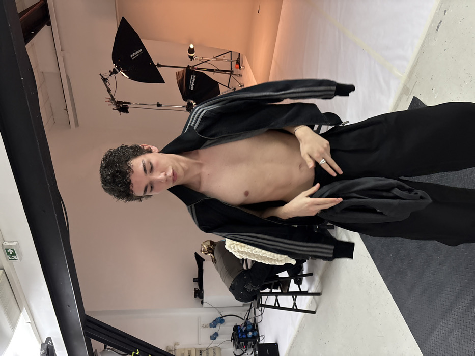 The image captures an indoor studio scene, located in Paris, France. A young, lean individual with dark, curly hair stands centrally, looking downwards with a neutral expression. They wear an open black track jacket with three light-colored stripes on the sleeves, exposing their bare chest and abdomen, paired with loose-fitting black trousers. A silver-colored ring adorns their right index finger, and their hands are positioned at waist level, holding a folded piece of dark fabric. In the left background, another individual is seated on a black wooden stool, their back partially to the camera. This person wears a brown long-sleeved top and dark trousers with light embellishments on the rear pockets, and appears to be tending to something near the floor. A light-colored, textured garment is draped over the stool. The studio features white walls, a high ceiling with an exposed black I-beam, and professional lighting equipment, including two Profoto softboxes on stands, positioned to the right. Electrical cables, power strips, and a Bose amplifier are visible on the floor on the left. A white seamless backdrop transitions to an orange hue under the studio lighting on the right. A green emergency exit sign is mounted on the wall in the upper left quadrant.