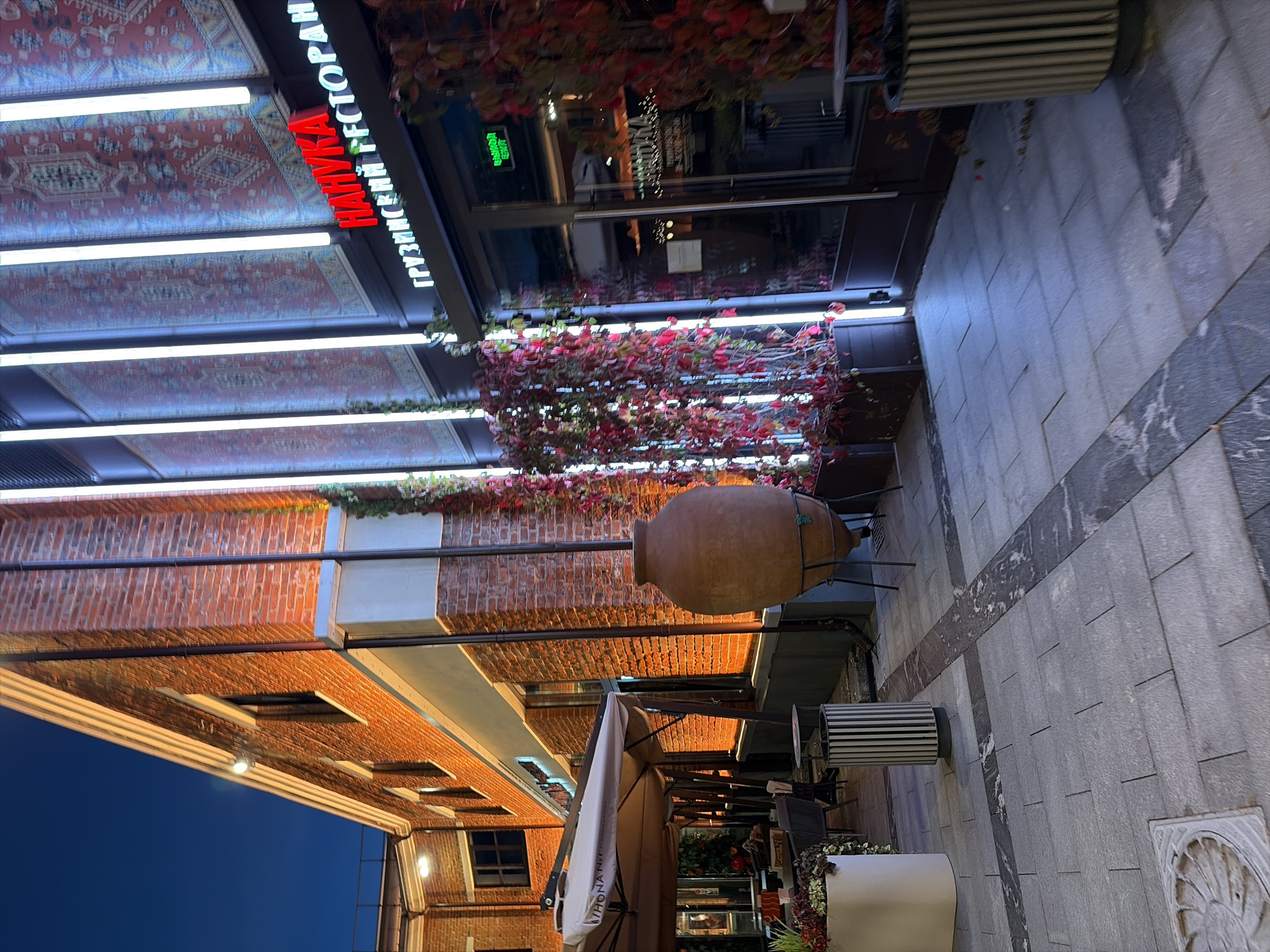 The image captures a street view at dusk in Moscow, Russia, outside a restaurant. The building facade is constructed of red brick and features vertical LED lights illuminating patterned carpets hung on the exterior wall. Above the entrance, a red sign reads "НАНУКА" (Nanuka), and below it, a green sign displays "ГРУЗИНСКИЙ РЕСТОРАН" (Georgian Restaurant). The entrance itself is a glass door. Red ivy plants cascade down the side of the building. In the foreground, a paved walkway is laid with grey rectangular stones and accented with darker stone inlays. A large, terracotta amphora stands on a metal stand to the right of the walkway. To the left, there are outdoor seating areas with tables and chairs, partially covered by a large umbrella. A cylindrical trash bin with vertical slats is positioned near the seating area. The overall atmosphere is illuminated by artificial lights, suggesting evening hours.
