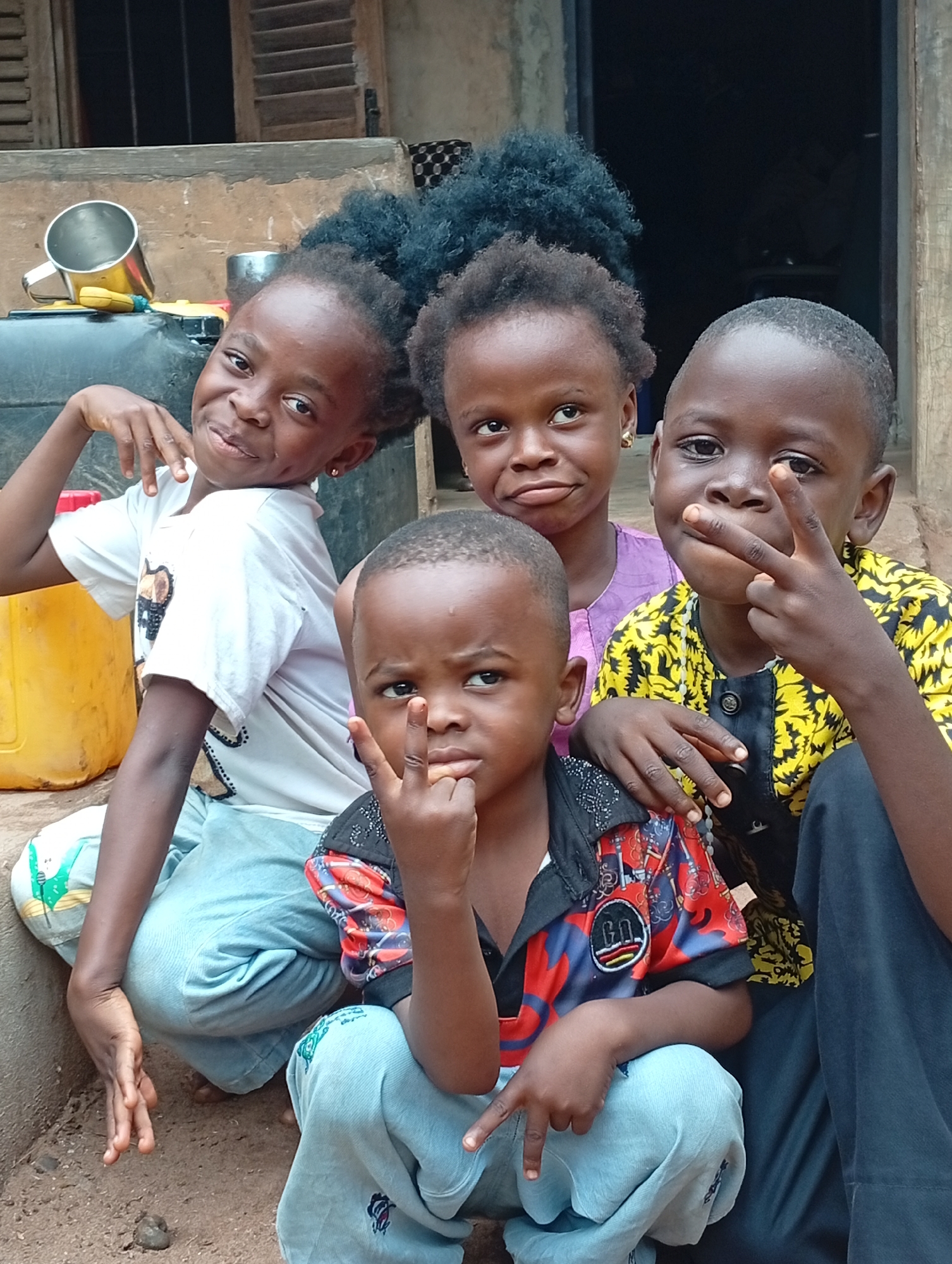 In Adda, Nigeria, four children are gathered outdoors, seated on a dirt ground in front of a building. The child on the far left, a girl, wears a white t-shirt and blue pants, striking a pose with her hand on her hip. Next to her, another girl in a purple top also looks towards the camera. The youngest child, a boy in a colorful shirt and blue pants, is in the foreground, making a peace sign with his fingers. To his right, a boy in a yellow and black patterned shirt and black pants also makes a peace sign. The background reveals the entrance of a building with wooden shutters and a textured wall. A large black water tank and a yellow plastic container are visible to the left of the children.