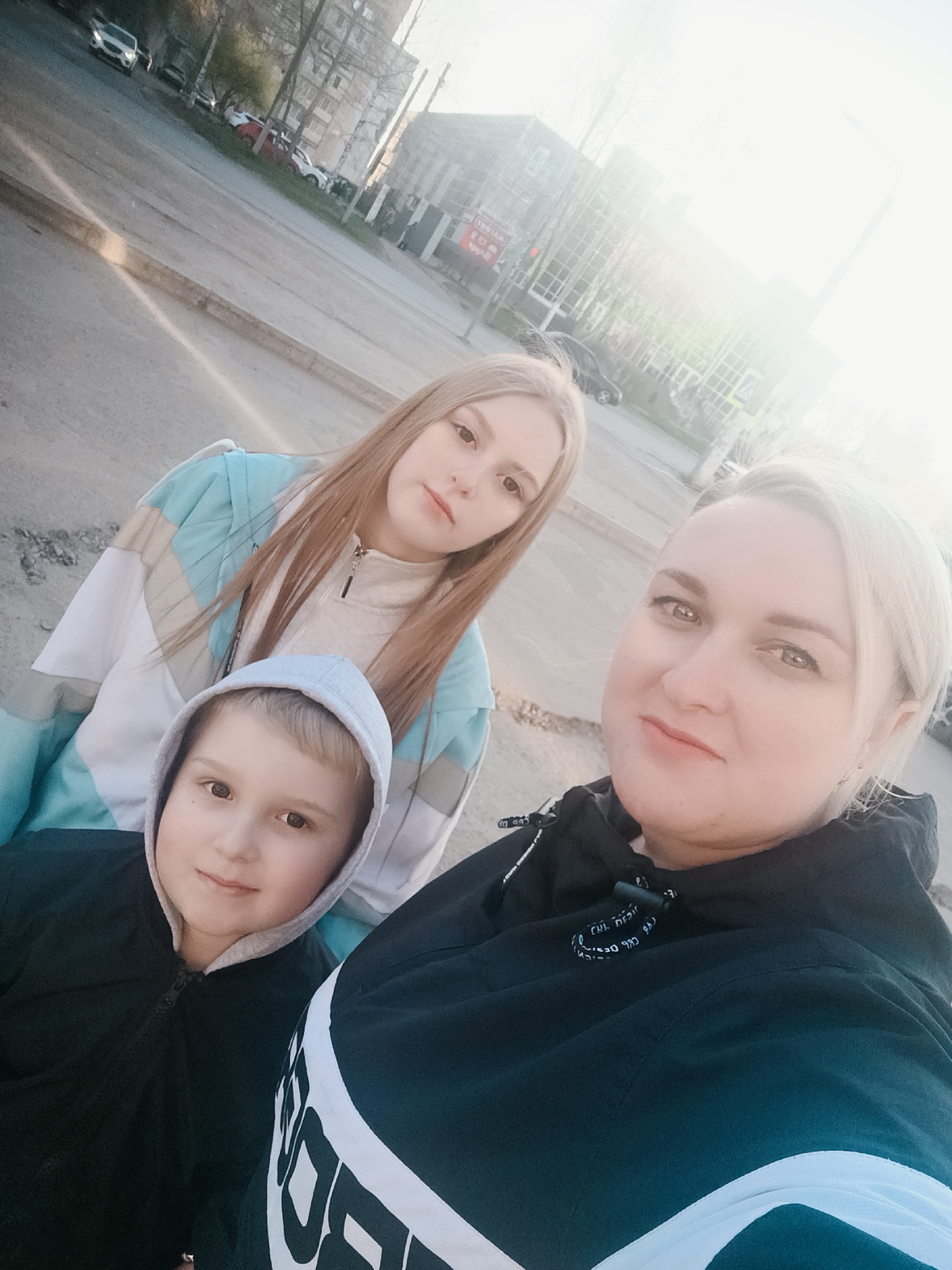 The photograph captures three individuals in an outdoor setting, likely in Chaikovsky, Russia, given the presence of typical Russian urban architecture in the background. A woman with blonde hair is holding the camera, positioning herself in the foreground. To her left stands a fair-haired young woman wearing a white top and a light blue, white, and beige jacket. In front of them is a young boy with a hooded sweatshirt. The backdrop includes a street and building with many windows. Several vehicles are parked along the street. The sky is bright and seems to be overcast. The ground appears to be asphalt.