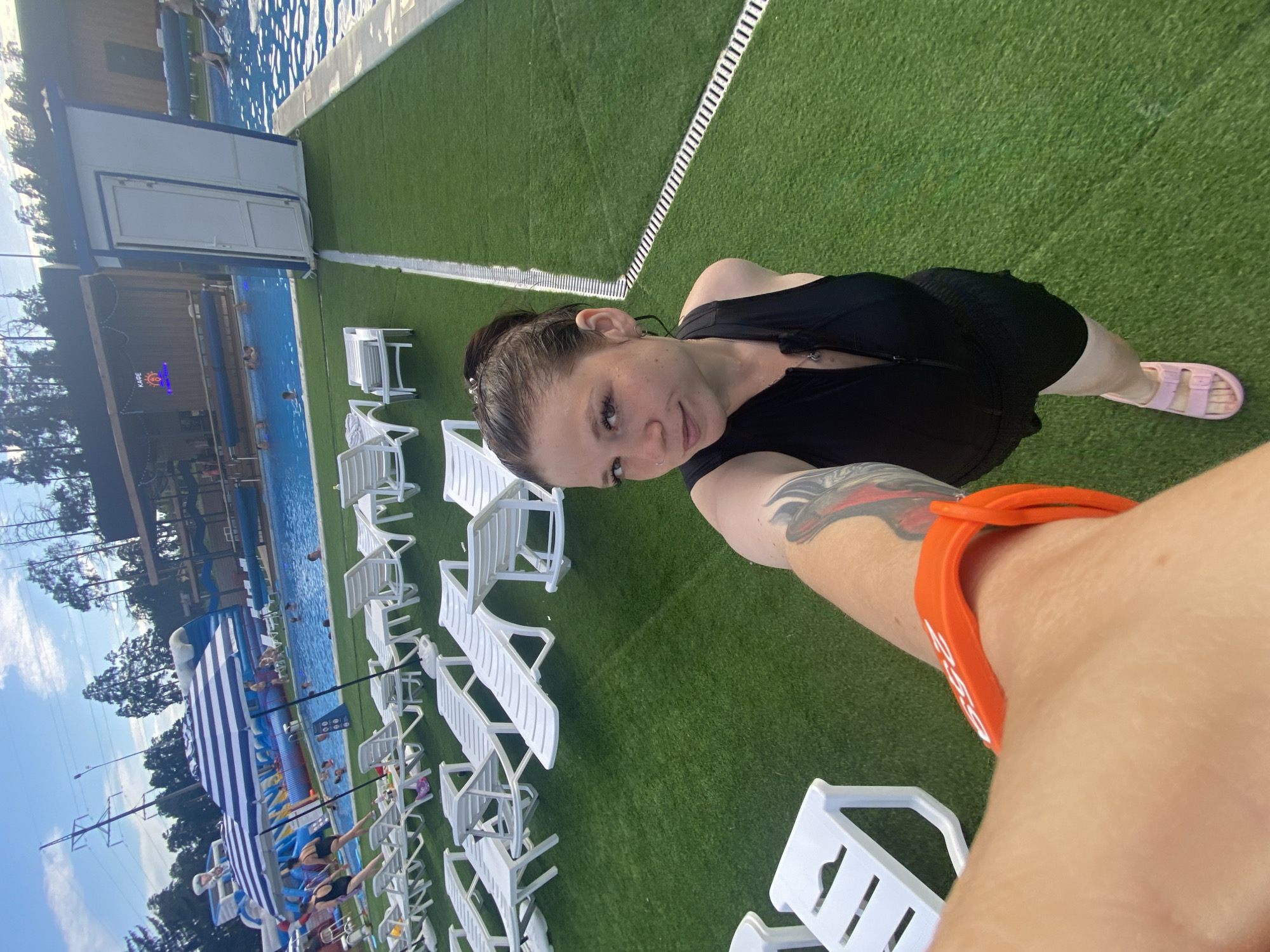 This image shows a woman taking a selfie at an outdoor water park in Votkinsk, Russia. The woman is wearing a black swimsuit with a zipper and pink sandals. She has an orange wristband on her left arm. In the background, there is a swimming pool with people swimming, lounge chairs, umbrellas, and a water slide. The ground is covered in artificial green grass. The sky is blue with some clouds. The overall scene depicts a leisure activity at a water park.