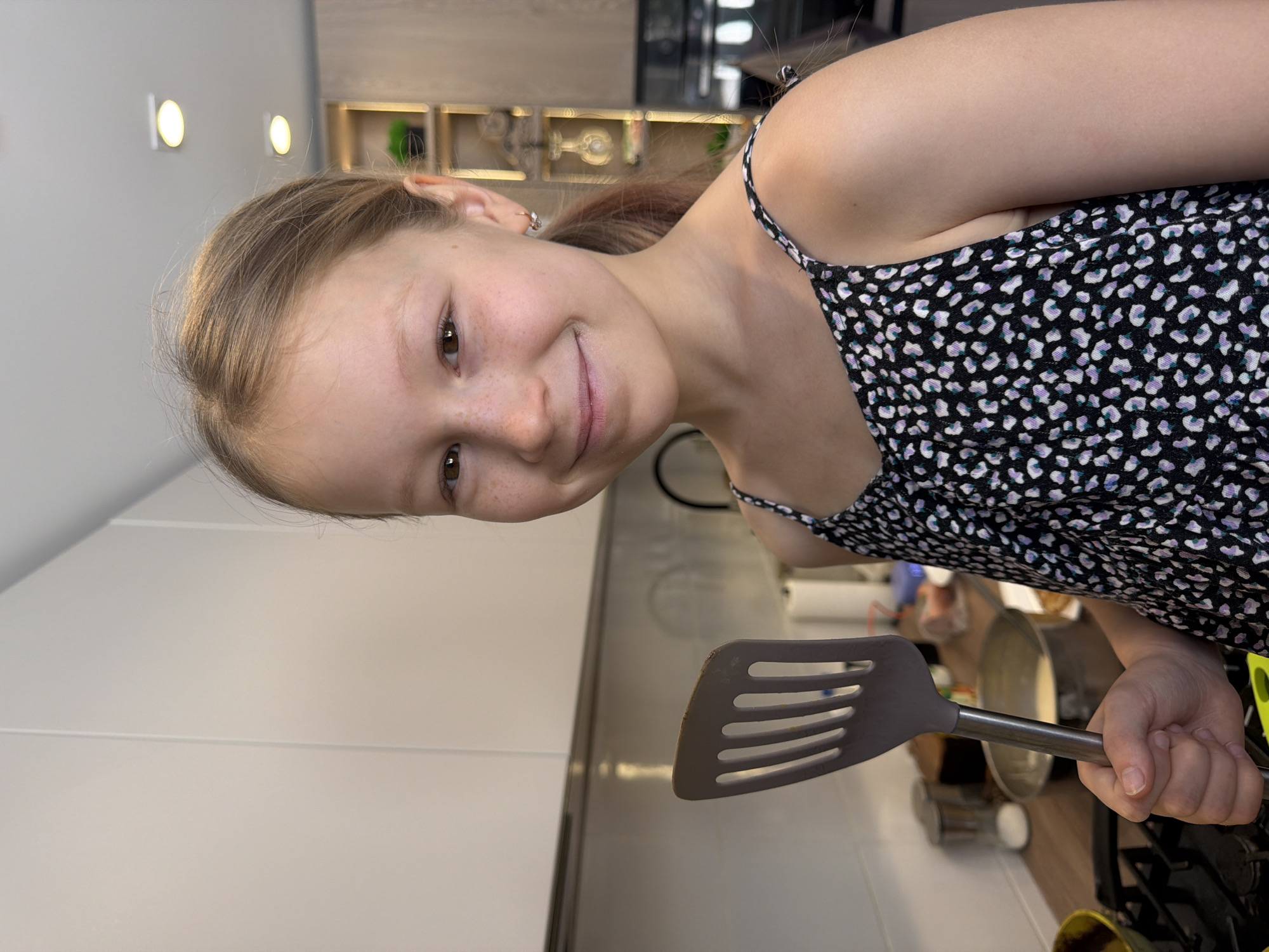 A young female subject is depicted in a residential kitchen in an unknown city. The subject, a child or pre-teen with light brown hair, fair skin, and freckles, is smiling directly at the camera. She wears a black camisole-style top with a white and light purple/blue irregular dot pattern and small silver hoop earrings. In her right hand, she holds a gray slotted spatula with a metallic handle. The background features a modern kitchen, including white overhead cabinets, a white tiled backsplash, and a light-colored countertop. A gas stovetop with black grates is visible in the lower left. On the countertop behind the subject, a metal bowl contains a light-colored liquid, alongside a clear jar with a white substance. Further in the background, a wooden shelving unit with a green potted plant and decorative items is visible, under recessed ceiling lights.