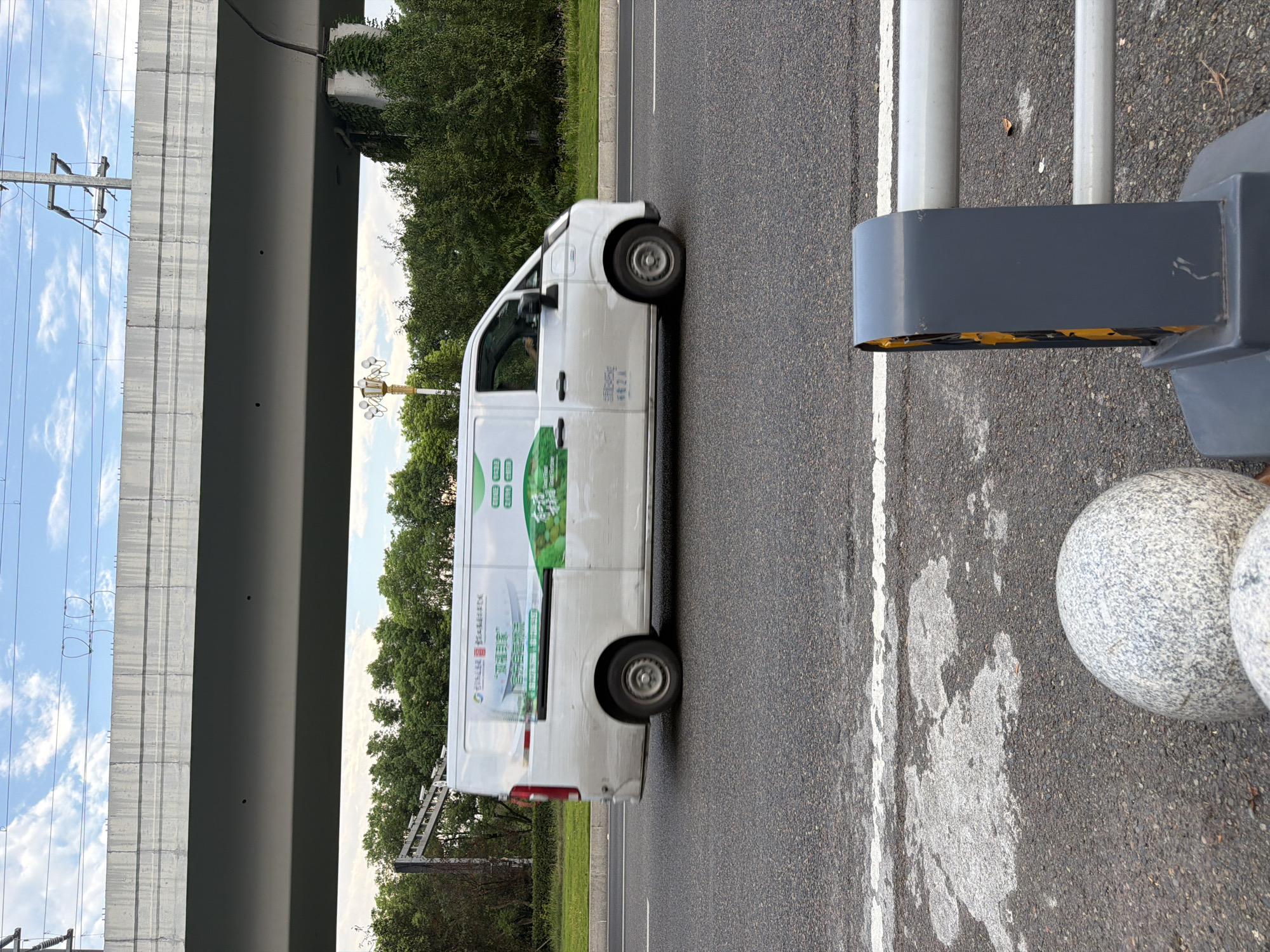 A white van with green graphics is driving on a road in Jiangjin, China. The van has Chinese text and images of green vegetables on its side. The road is asphalt and has white lane markings. In the background, there are trees, a highway overpass, and power lines. The sky is cloudy. In the foreground, there are two large, round, grey stone bollards and a traffic barrier.