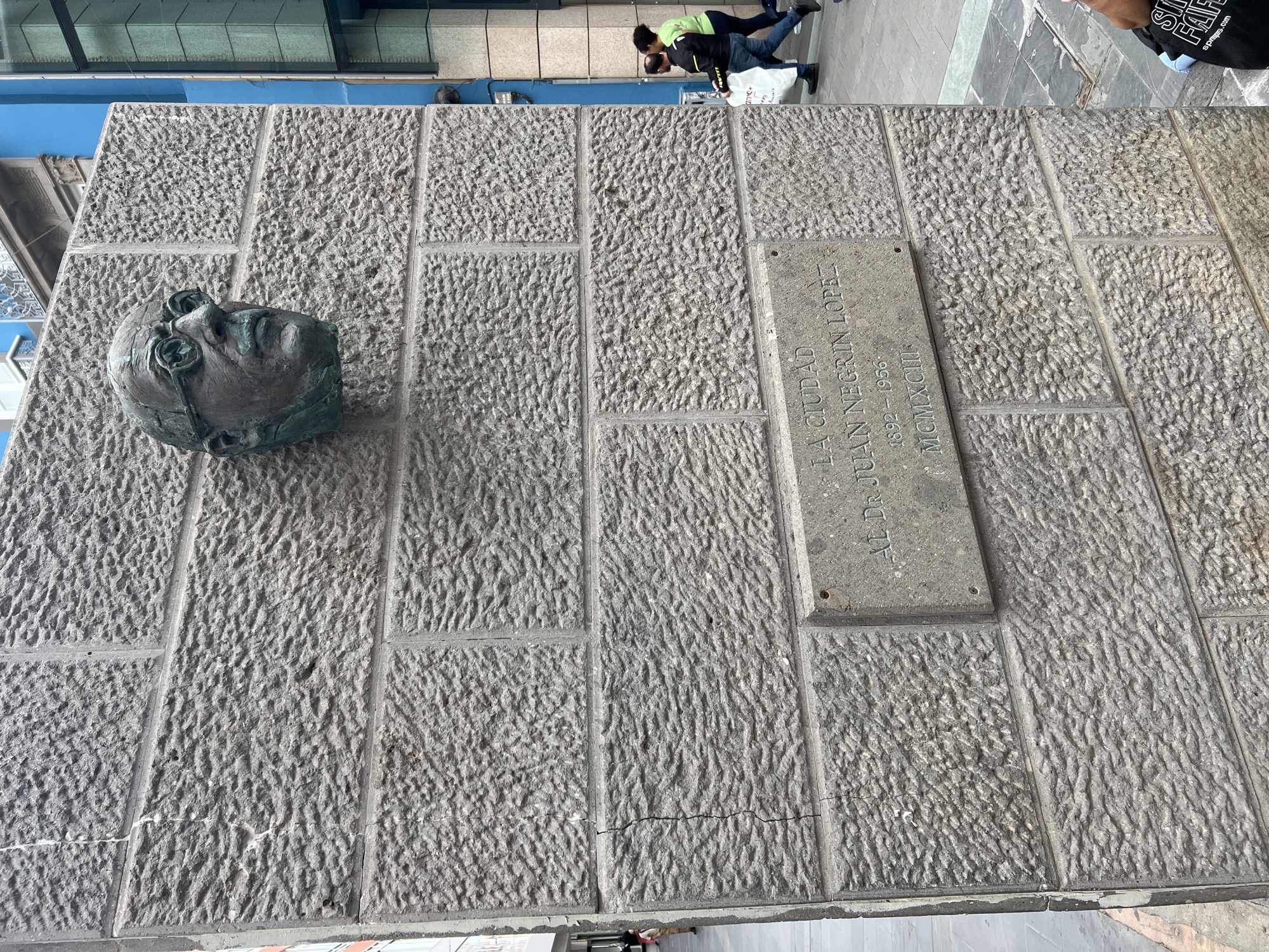The image shows a stone wall in Las Palmas De Gran Canaria, Spain. A bronze head is mounted on the left side of the wall. To the right of the head, a plaque reads "LA CIUDAD AL Dr. JUAN NEGRIN LOPEZ 1892-1956 MCMXCIII". Two people are visible in the background. One is wearing a green jacket. A third person is partially visible in the upper right corner.