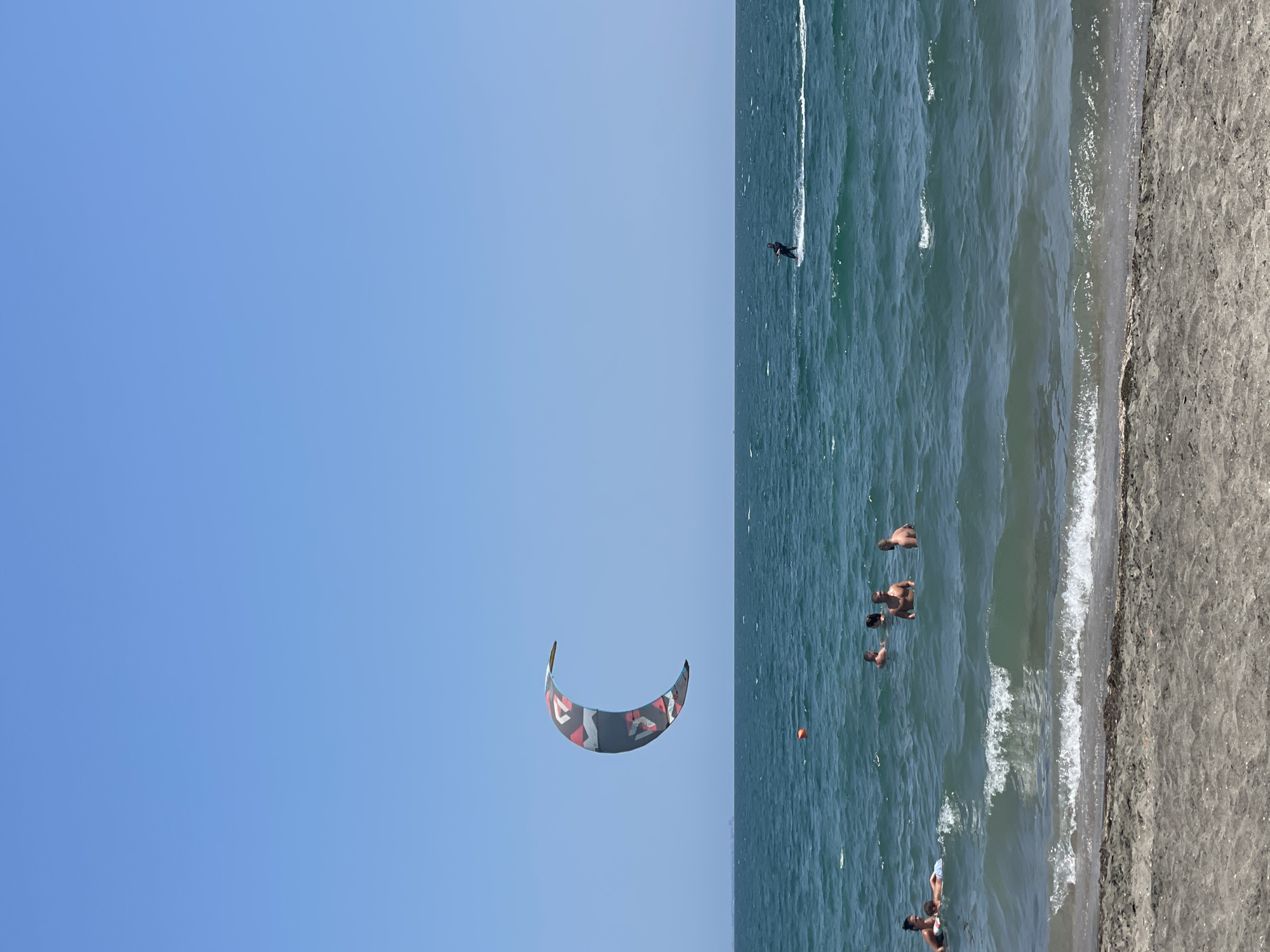 The image depicts a sunny day at a beach, likely on the coast of Burgas, Bulgaria. In the foreground, a sandy beach meets the choppy, teal-colored sea. Several people are in the water, some appearing to be swimming or wading, while others are further out, engaged in water sports. A kitesurfer is visible in the middle ground, riding on the water's surface. Overhead, a large, crescent-shaped kite, patterned with red, white, and black geometric shapes, is suspended in the bright blue sky. The horizon is visible in the distance, with a faint line of buildings suggesting a coastal town or city. An orange buoy floats in the water closer to the shore.