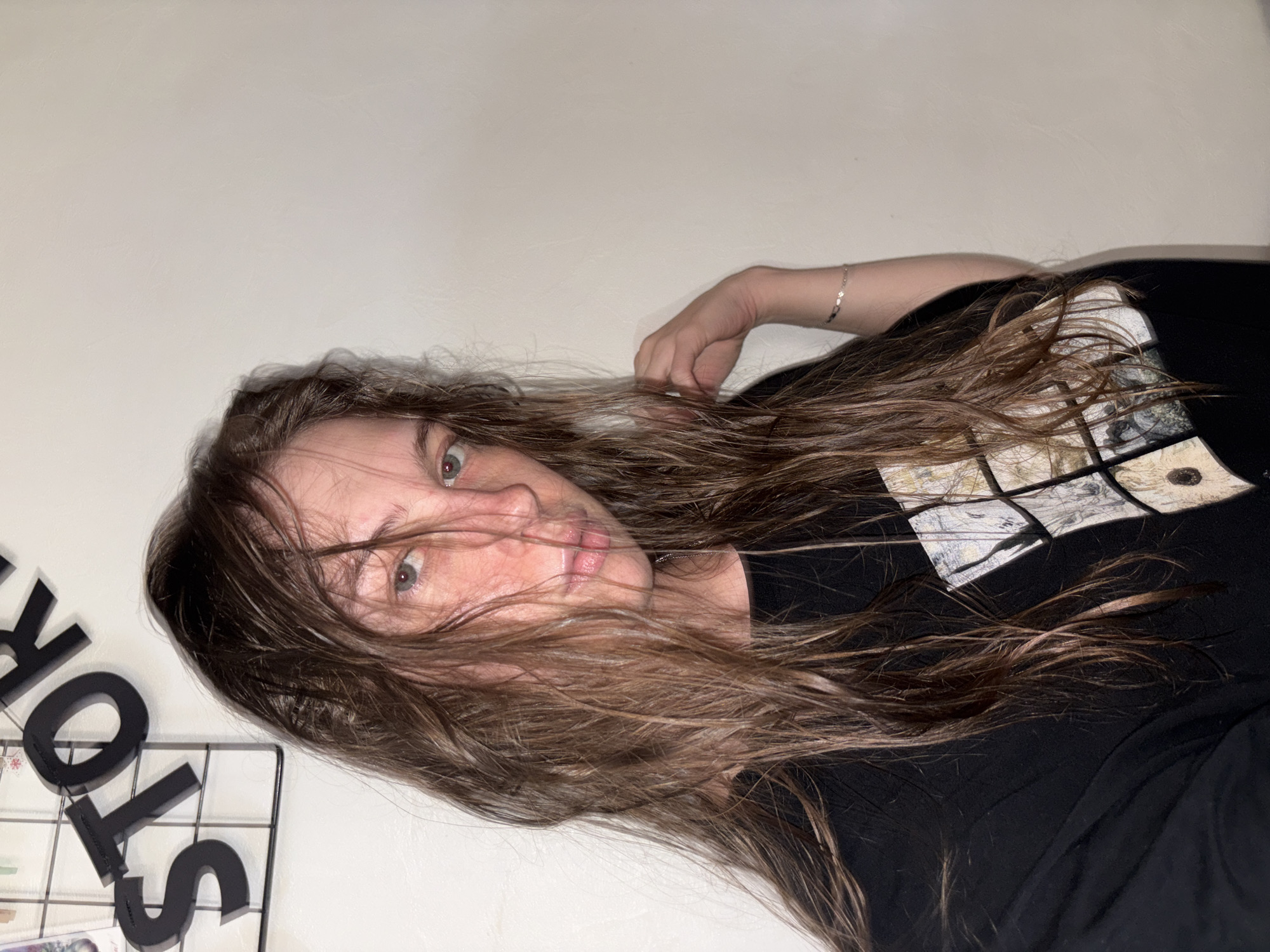 The image shows a person with long, brown hair lying down. Their face is partially obscured by their hair, but their eyes are visible and looking towards the camera. They are wearing a black t-shirt with a graphic print. On the left side of the image, there are black letter cutouts on a wire grid, spelling out "STORY". The background is a plain, light-colored wall. The lighting suggests an indoor setting, possibly at night, with a direct light source creating shadows. The person appears relaxed, with their hand resting on their hair. There are no obvious indications of the weather or specific location beyond the presence of the letters, which could be part of a decorative element or a personal project. The overall mood is casual and intimate.