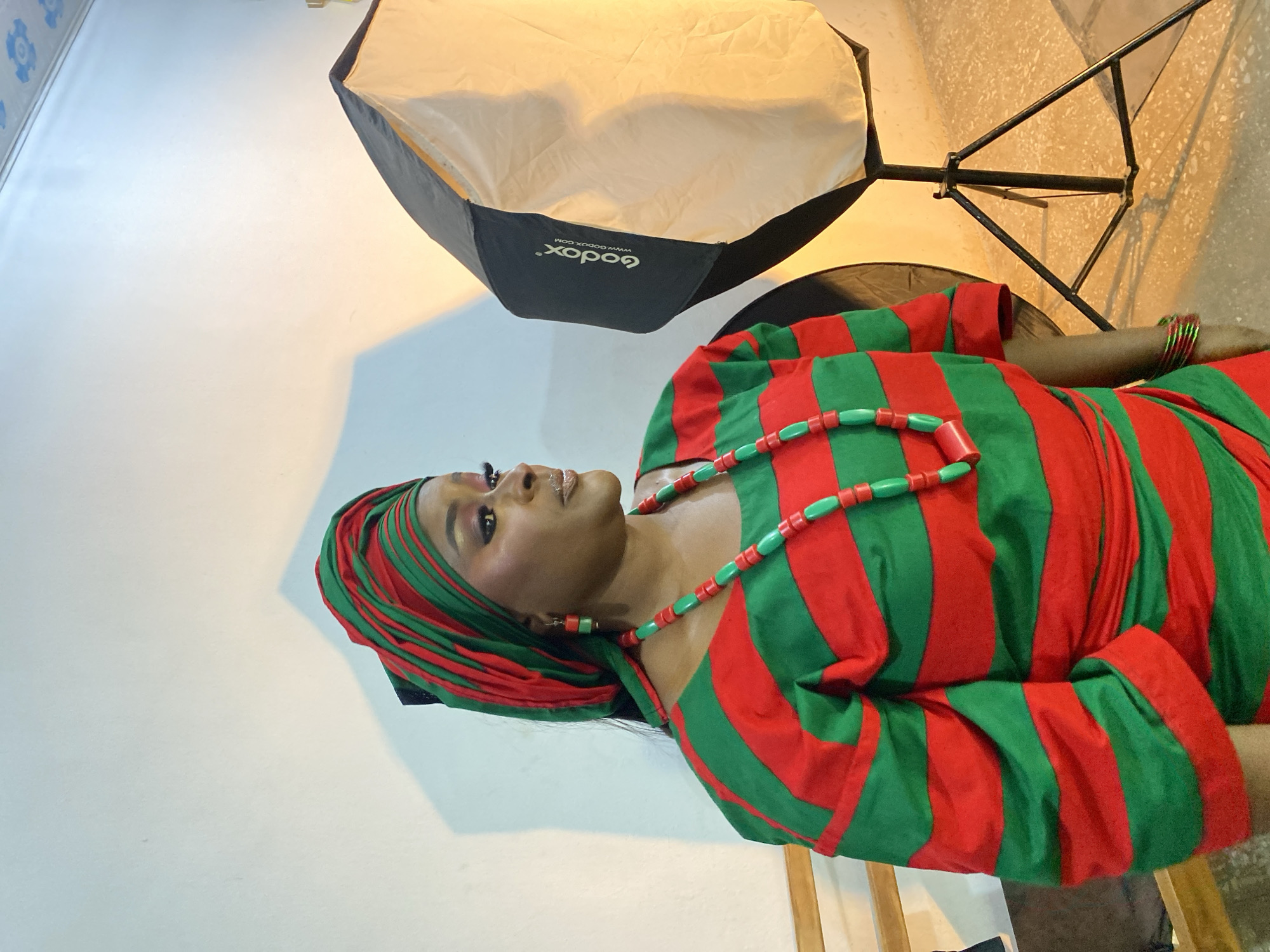 A medium shot depicts a woman in traditional Nigerian attire and jewelry standing in front of a studio backdrop. She is wearing a red and green striped dress with a matching headwrap and a necklace made of red and green beads. Her makeup is done with gold eyeshadow, and she is wearing earrings that match her necklace. She is turned to the left and looking up. Next to her is a softbox with the brand name "Godox" printed on it, mounted on a black metal stand. The backdrop is a plain white wall. The location is likely a photography studio in Gura Top, Nigeria.