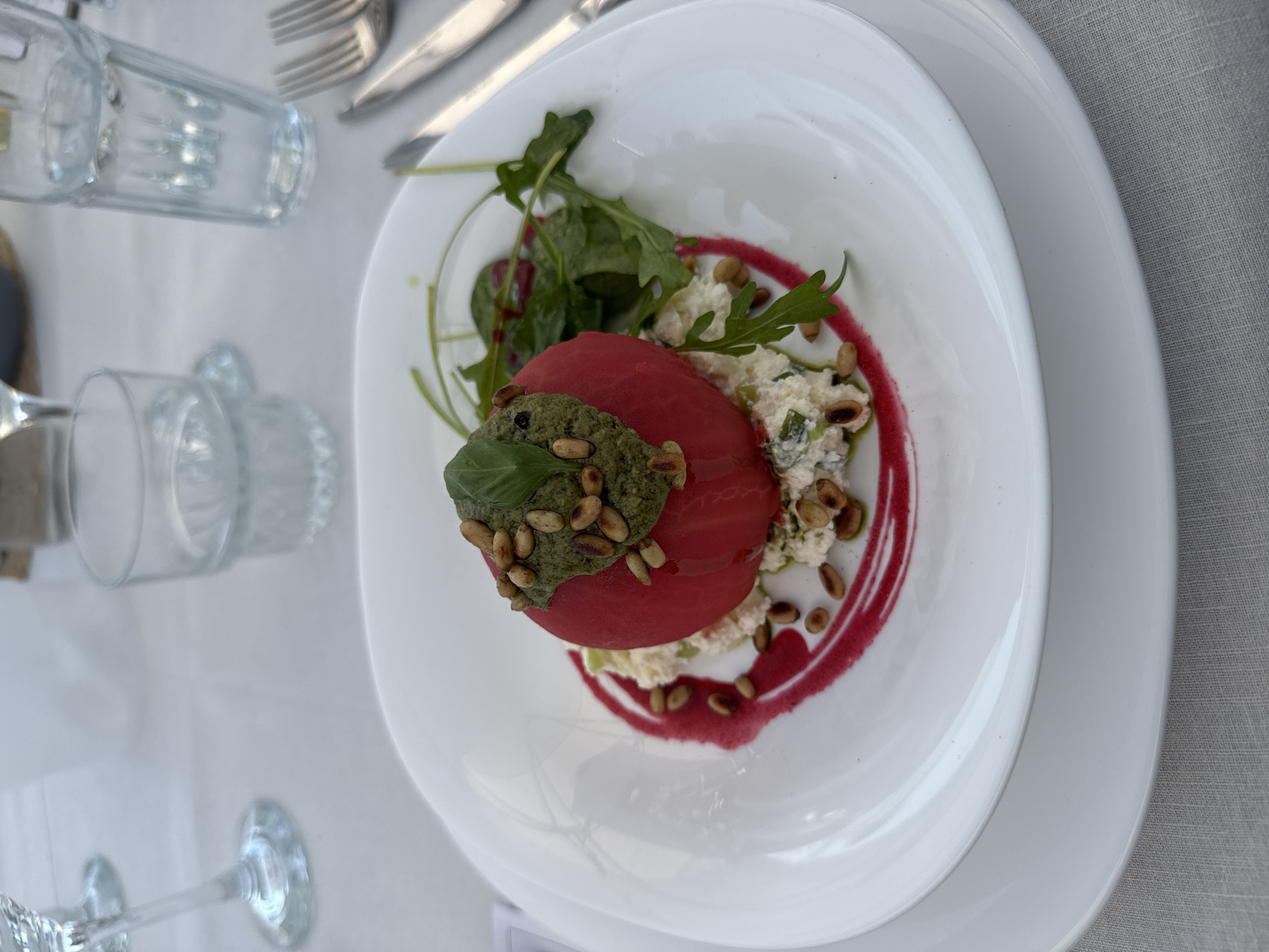 A high-angle, close-up photograph captures a meticulously plated culinary dish on a dining table in Harsovo, Bulgaria. The central subject is a vibrant red, peeled tomato, possibly roasted or blanched, resting on a bed of a textured, white, creamy substance resembling a cheese mixture, which contains finely chopped green elements. The tomato is crowned with a generous portion of dark green pesto, adorned with numerous toasted pine nuts and a single fresh basil leaf. A deep red liquid, possibly a berry or beetroot puree, is artistically swirled in a crescent shape beneath the white mixture, extending from the bottom left towards the center. Several individual pine nuts are scattered around the plate, resting on both the white mixture and the red swirl. To the right of the central dish, a cluster of fresh green leaves, likely arugula, adds a fresh garnish. The dish is presented on a white, round plate with a subtle rim, which is placed atop a larger, also white, round underplate. The table is covered with a light grey, textured fabric tablecloth. In the blurred background, several clear drinking glasses are visible: a stemmed glass in the upper left, and two water glasses, one partially obscured, containing clear liquid, in the upper right. Silver cutlery, consisting of a fork and knife, is partially visible on the right side of the plate. No human subjects are present in the frame.