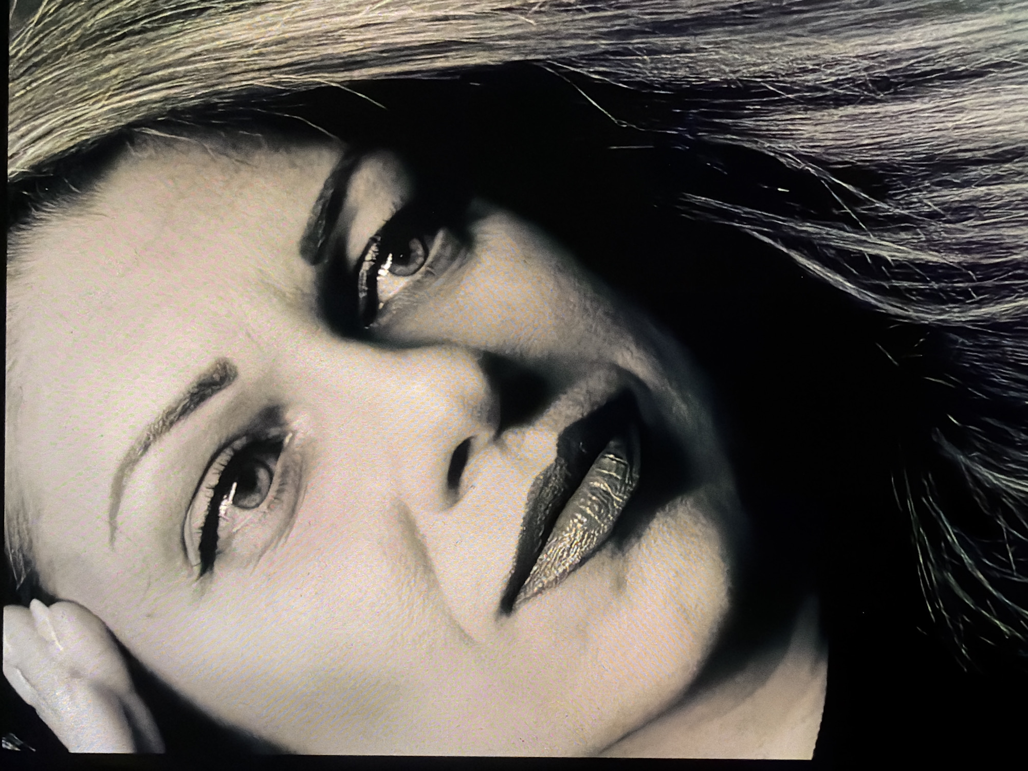 A close-up, monochrome image shows a woman's face. She is fair-skinned with dark eyes and light, streaked hair that is partially obscuring her face. Her head is tilted slightly to the right, with her left hand visible near her cheek. Her lips appear to have some texture and are closed. The lighting is uneven, creating strong shadows on the right side of her face and hair. The photograph lacks specific details connecting it to Dryanovo, Bulgaria. Therefore, without additional information, no location within the city can be determined.