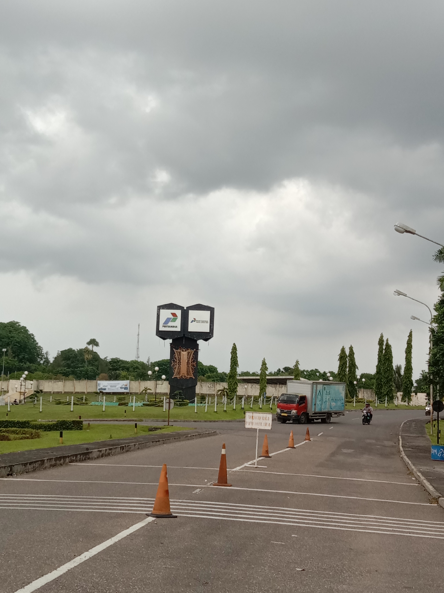 The image depicts a wide paved road leading towards a monument with a Pertamina logo in Lhokseumawe, Indonesia. Several orange traffic cones are placed across the road. A red truck with a white cargo container passes by. A motorcyclist rides on the right side of the road. The sky is overcast. Green lawns and trees flank the road, with a white wall in the background. Streetlights are visible along the road. A sign with Indonesian text stands near the traffic cones.