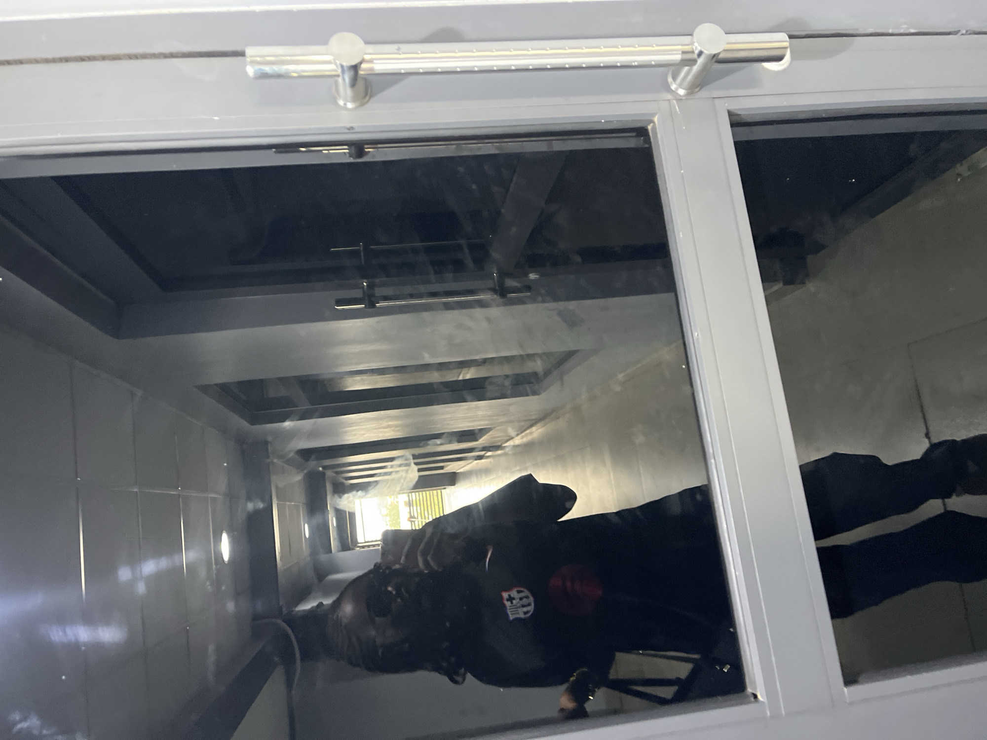 A person is reflected in a glass window. The window appears to be part of a structure with a repetitive, tunnel-like design. The walls of this tunnel are tiled and feature evenly spaced light fixtures. The person is wearing dark clothing with a badge on the chest. The location is Karum, Nigeria.
