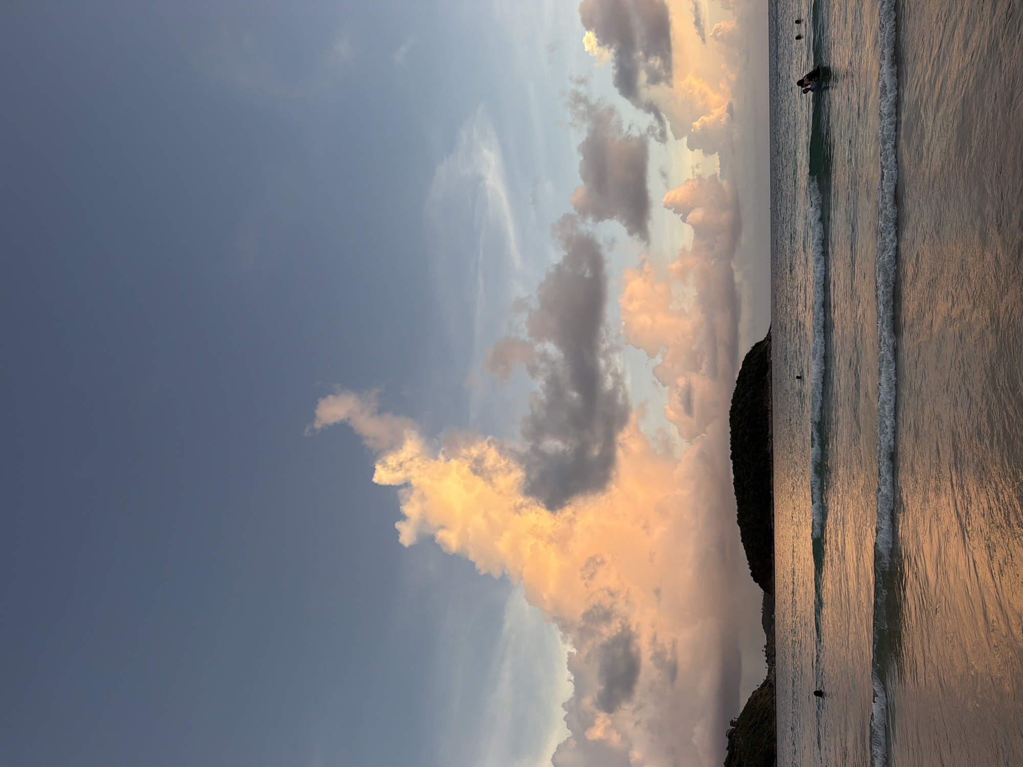 The image depicts a serene sunset scene at a beach in Rawai, Thailand. The sky is painted with hues of blue, orange, and pink, with dramatic clouds illuminated by the setting sun. In the foreground, gentle waves lap the sandy shore, reflecting the warm colors of the sky. The ocean stretches out to the horizon, where two islands covered in lush green vegetation are visible. Several people are scattered across the water, some swimming and others wading. One person appears to be on a surfboard, while others are simply enjoying the calm sea. The overall atmosphere is tranquil and picturesque, capturing the beauty of a tropical evening.