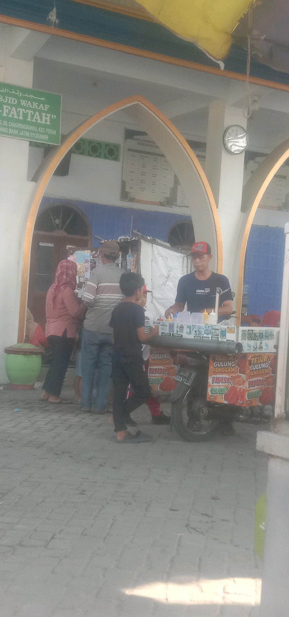 In Temuwulan, Indonesia, a street scene unfolds near the Masjid Waqaf Fattah. The mosque features a prominent arched entryway. In the foreground, a mobile food cart, likely selling local snacks like "Telur Gulung" (rolled eggs) and "Papeda" (a starchy dish), is a central element. The cart is decorated with colorful signage advertising these items. A man wearing a black t-shirt and a red cap stands behind the cart, appearing to serve customers. Several individuals are gathered around the cart, including a woman in a pink hijab and patterned tunic, a man in a striped shirt, and two boys. One boy, in a black t-shirt, stands facing the cart, while another, wearing a red cap, is partially visible. The ground is paved with light-colored bricks. A green pot and a clock on a pillar are also visible within the scene, suggesting an outdoor setting. The overall impression is a typical street-level transaction in a community setting.
