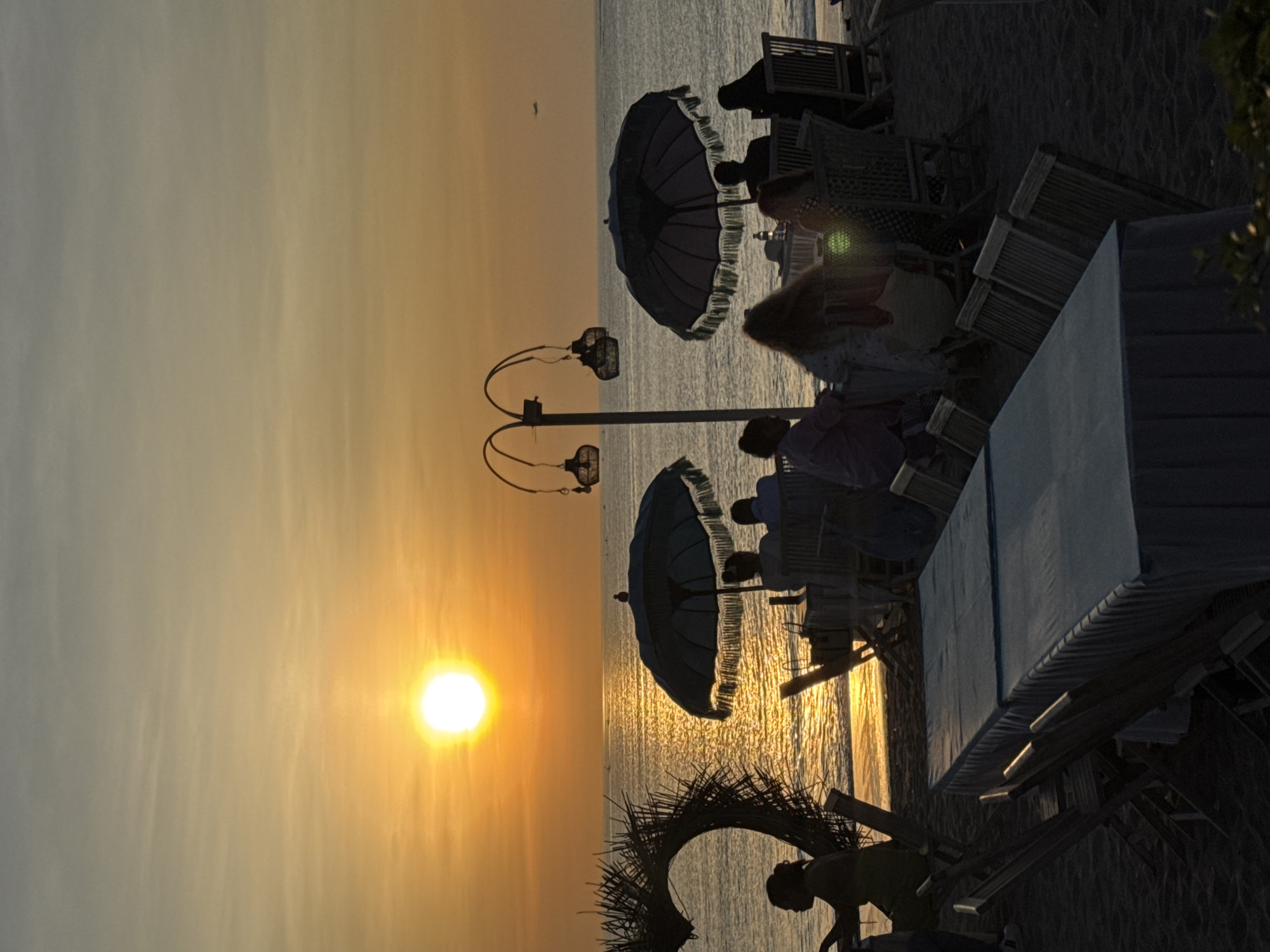 The image captures a sunset scene in Kuta, Indonesia, featuring a beach setting. The sun is low in the sky, casting a warm golden light over the ocean. Silhouetted figures of people are seated on beach chairs under umbrellas. A decorative arch made of natural materials is visible in the foreground. There is a lamppost with ornate lamps visible. The beach appears to be sandy, and the ocean extends to the horizon. The overall ambiance is peaceful and scenic.