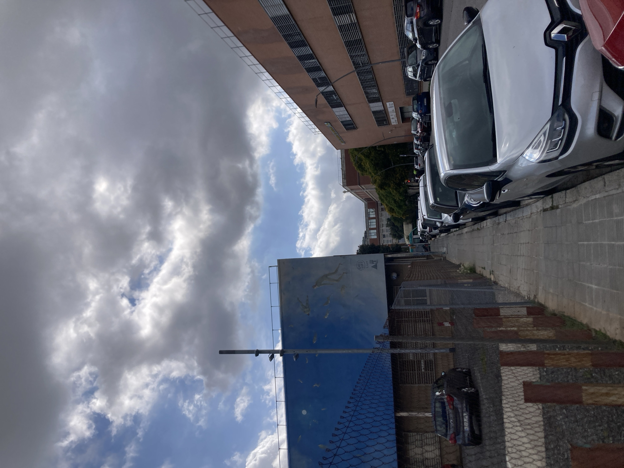 The image depicts a street scene in Barcelona, Spain, on a cloudy day. In the foreground, a row of parked cars lines the right side of the street. The cars vary in color and model, with a silver Renault Clio prominently visible. The left side of the street features a blue building with a mural depicting figures in water, possibly a swimming pool or ocean scene. A chain-link fence separates the sidewalk from a gravel area next to the blue building. Further down the street, a large, reddish-brown building with multiple rows of windows is visible. The sky is partly cloudy, with large formations of grey and white clouds. There are no visible people actively engaged in the scene. The environment appears to be an urban setting with a mix of residential and commercial or institutional architecture.