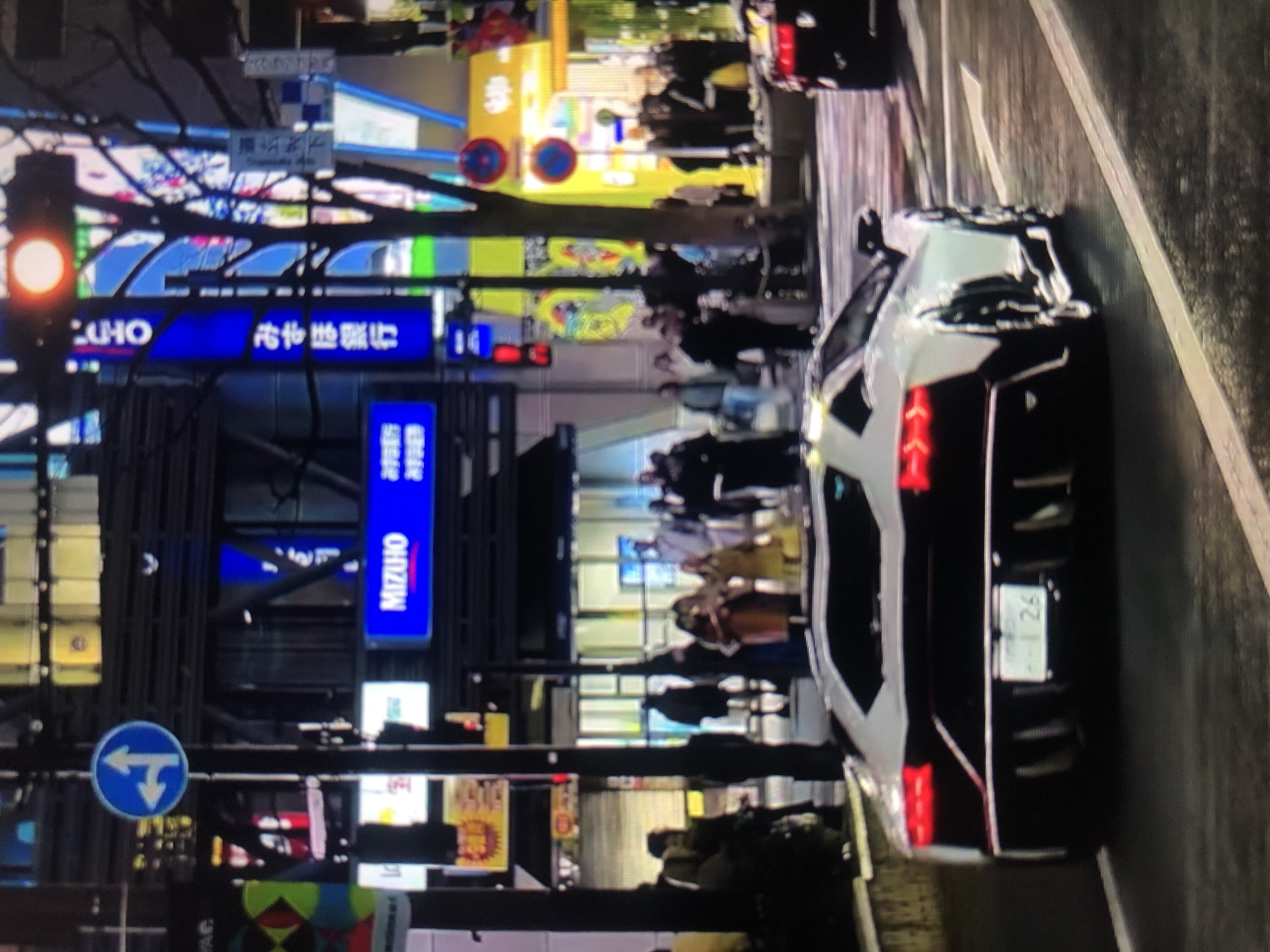 The image depicts an urban street scene at night, likely in Japan based on signage. A white and black sports car is parked on the right side of the frame. Pedestrians are visible walking on the sidewalk. Streetlights and neon signs illuminate the area, including signs for "MIZUHO" and "UCHO". A traffic light on the left side of the frame is showing red. A blue road sign with white arrows indicating left and downward directions is also visible.
There is no evidence that the image is in the city of Lhokseumawe, Indonesia.
